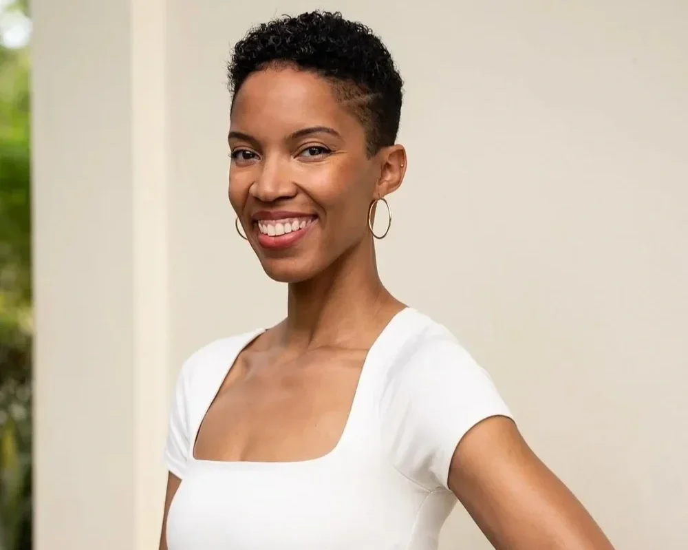 A woman with short curly hair smiling, wearing a white top and hoop earrings, standing outdoors near a light-colored wall.