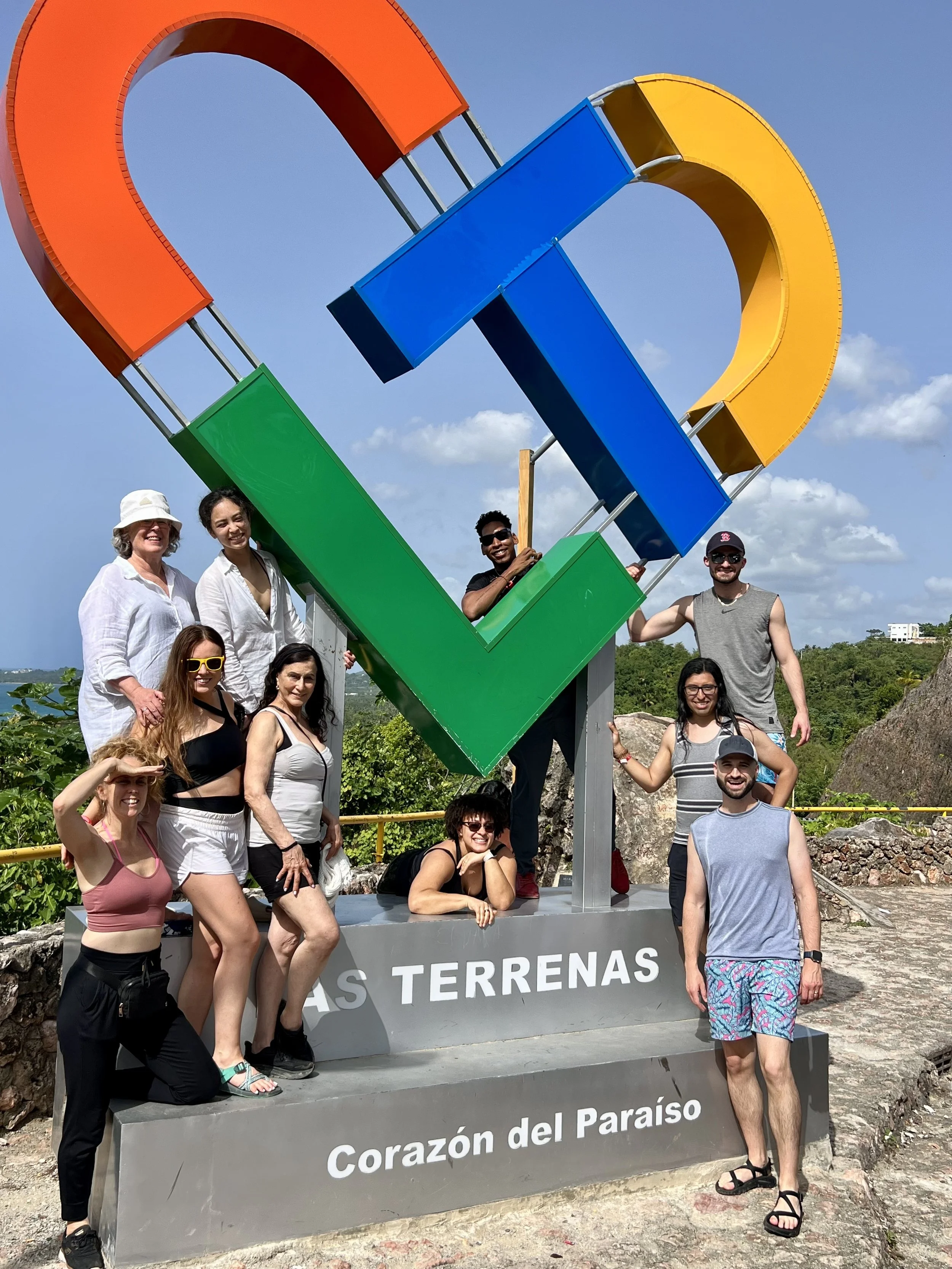 A diverse group of people posing around a large colorful sign that reads 'Corazón del Paraíso' and 'LAS TERRAZAS' with a scenic background of greenery, blue sky, and some buildings.