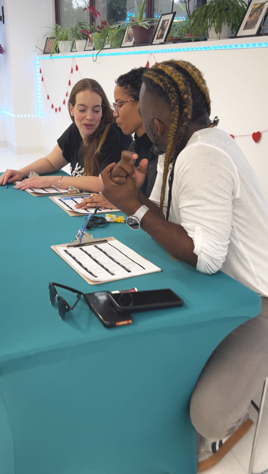 Three people sitting at a teal table, engaged in conversation, with clipboards, smartphones, sunglasses, and small items on the table, decorated with red hearts and string lights.