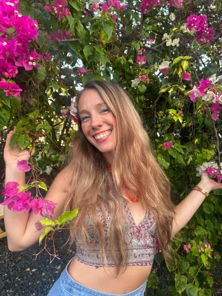 A young woman smiling in front of green foliage and pink bougainvillea flowers.