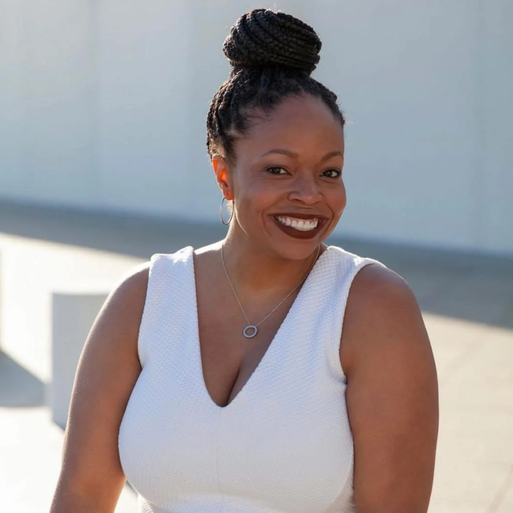 A woman smiling outdoors, wearing a white sleeveless top, hoop earrings, and a circular pendant necklace, with her hair styled in braids wrapped into a bun.