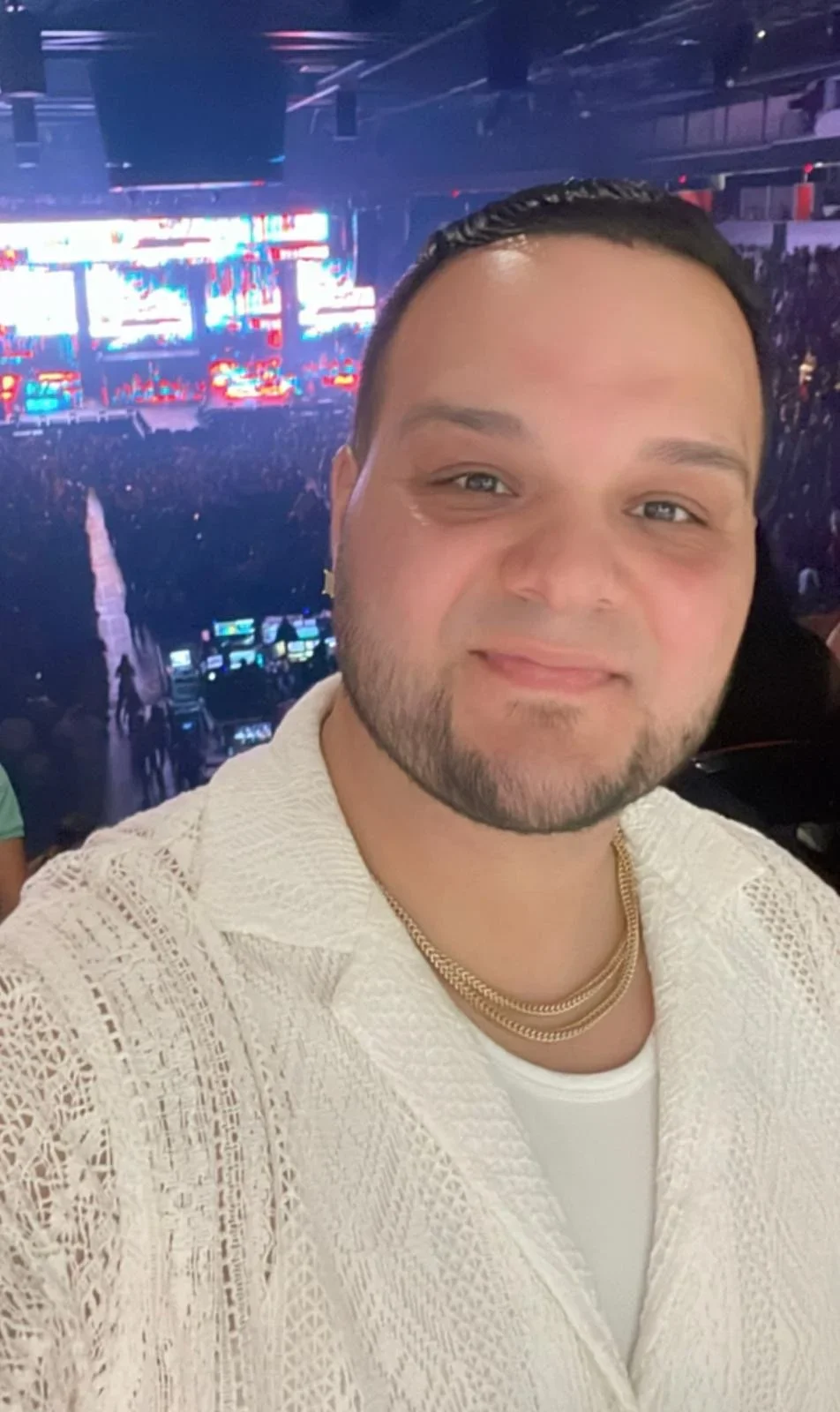 A man taking a selfie at a concert or event, with a large crowd and bright stage lights and screens in the background.