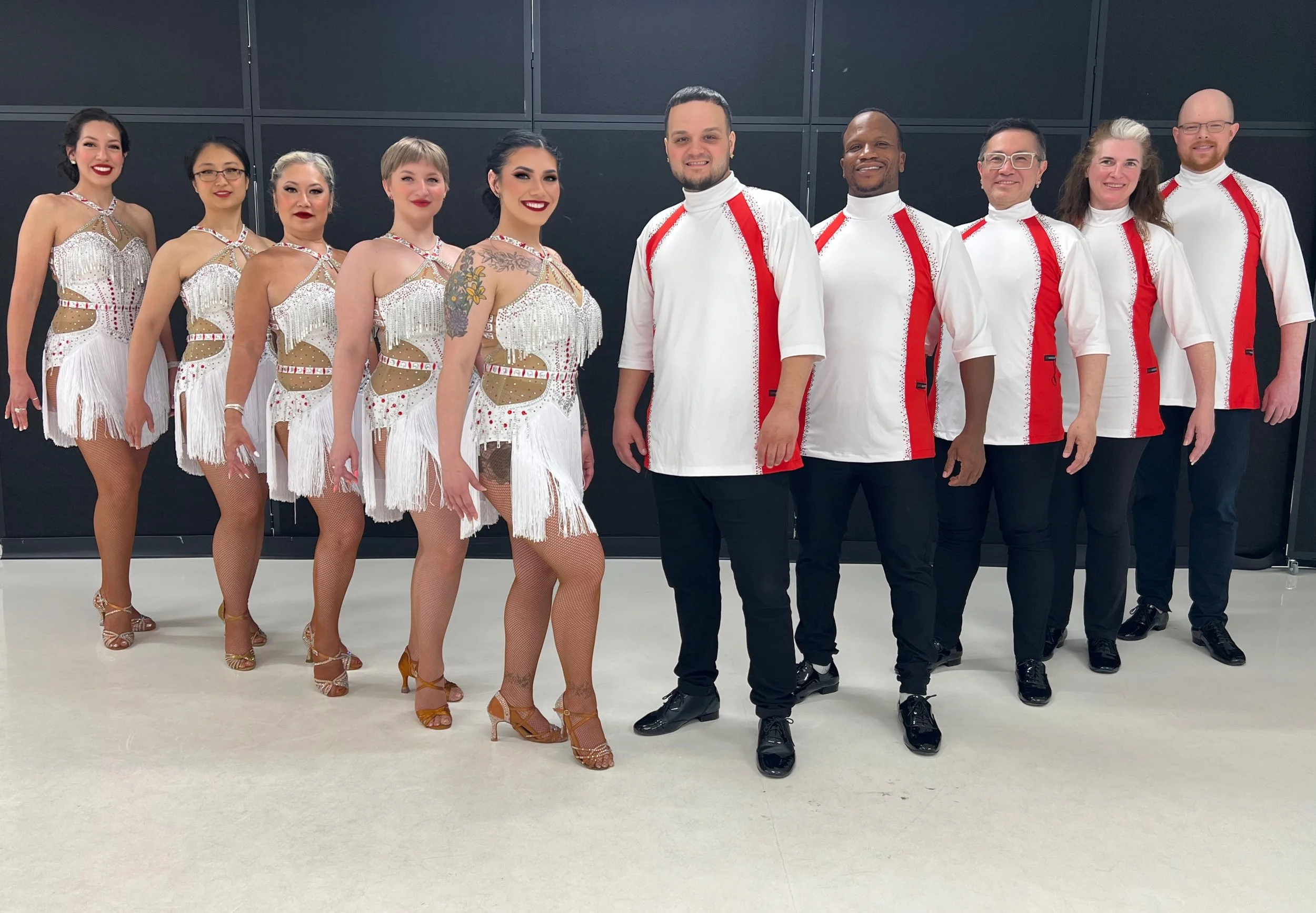 Group of ten dancers, six women and four men, standing in a line inside a room with a black wall background. The women are wearing sparkly, beige and white costumes with fringe, fishnet stockings, and dance shoes. The men are wearing white shirts with red accents and black shoes. The dancers are smiling and facing the camera.