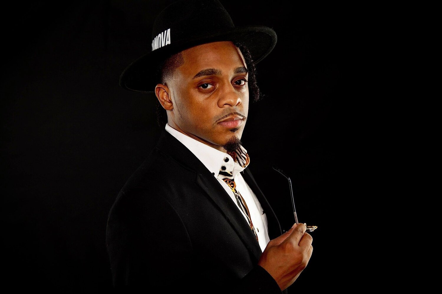 A man in a black suit and white shirt holding eyeglasses, wearing a black fedora hat with white text, against a black background.