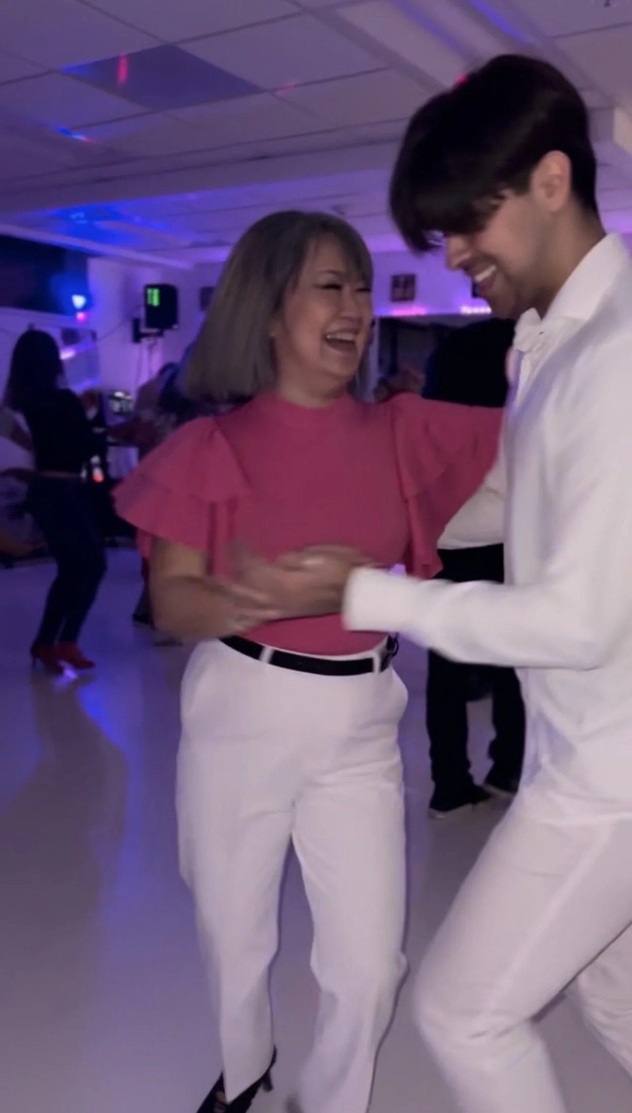 Two people dancing closely, both smiling and wearing white pants, with colorful lights and other people dancing in the background.