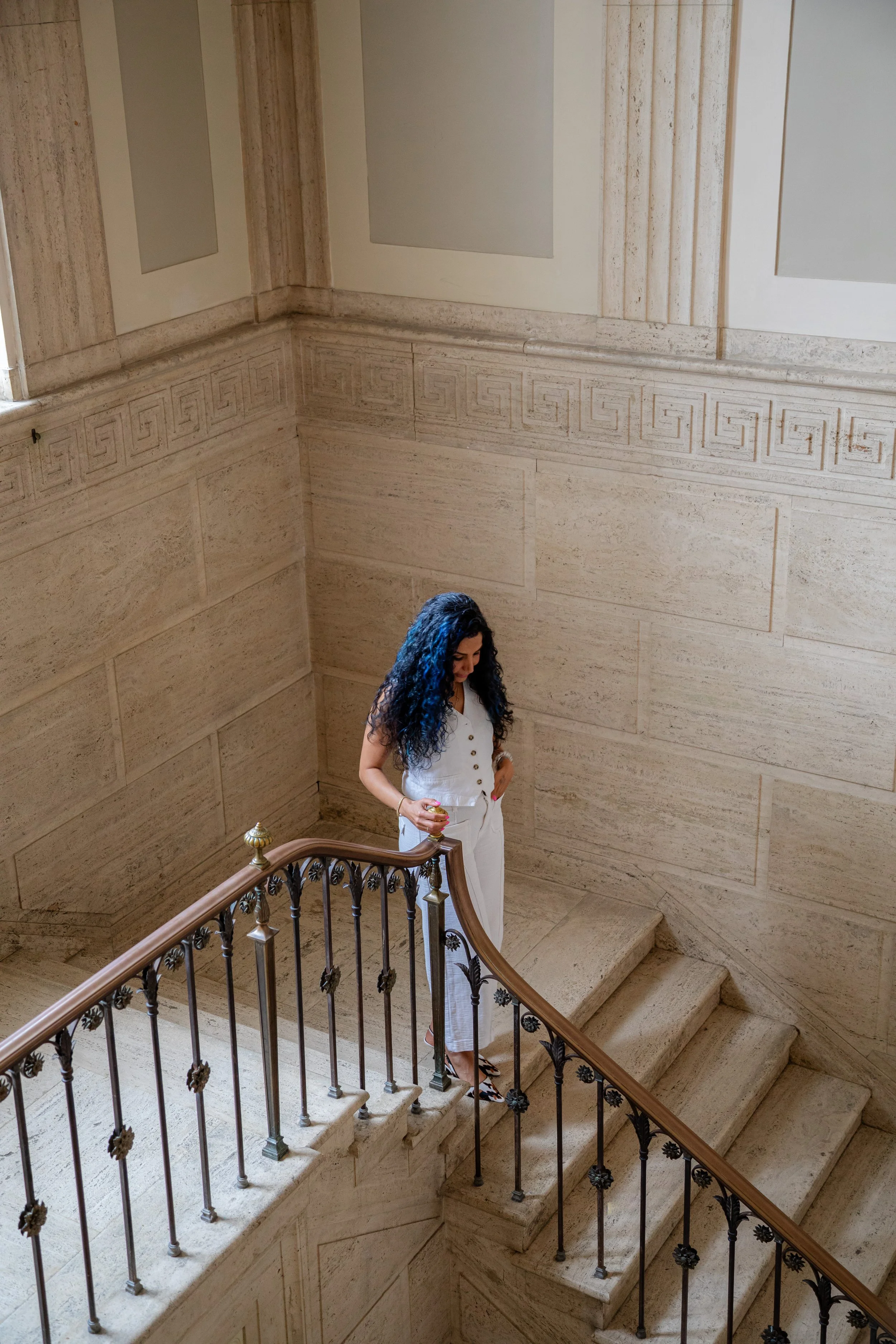 A woman with curly dark blue hair, wearing a white sleeveless top and white pants, stands on a staircase with ornate black metal railing and gold accents. She is holding a small object in her left hand and looking down. The staircase leads down to a beige stone hallway with decorative Greek key patterned wall border and large beige stone panels.