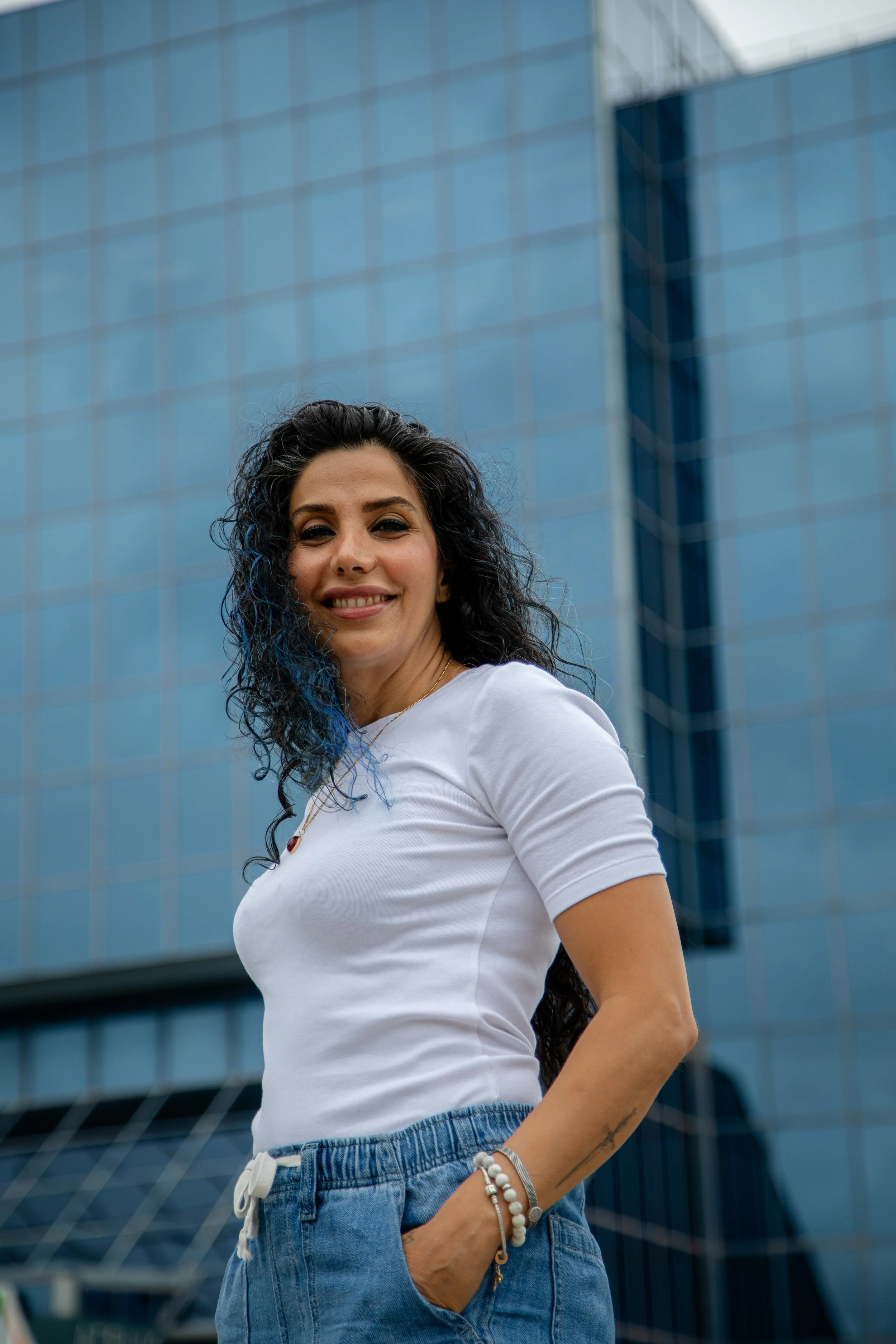 A woman with long, curly black hair and a nose piercing smiling in front of a modern glass building, wearing a white t-shirt and blue jeans, accessorized with bracelets and a necklace.