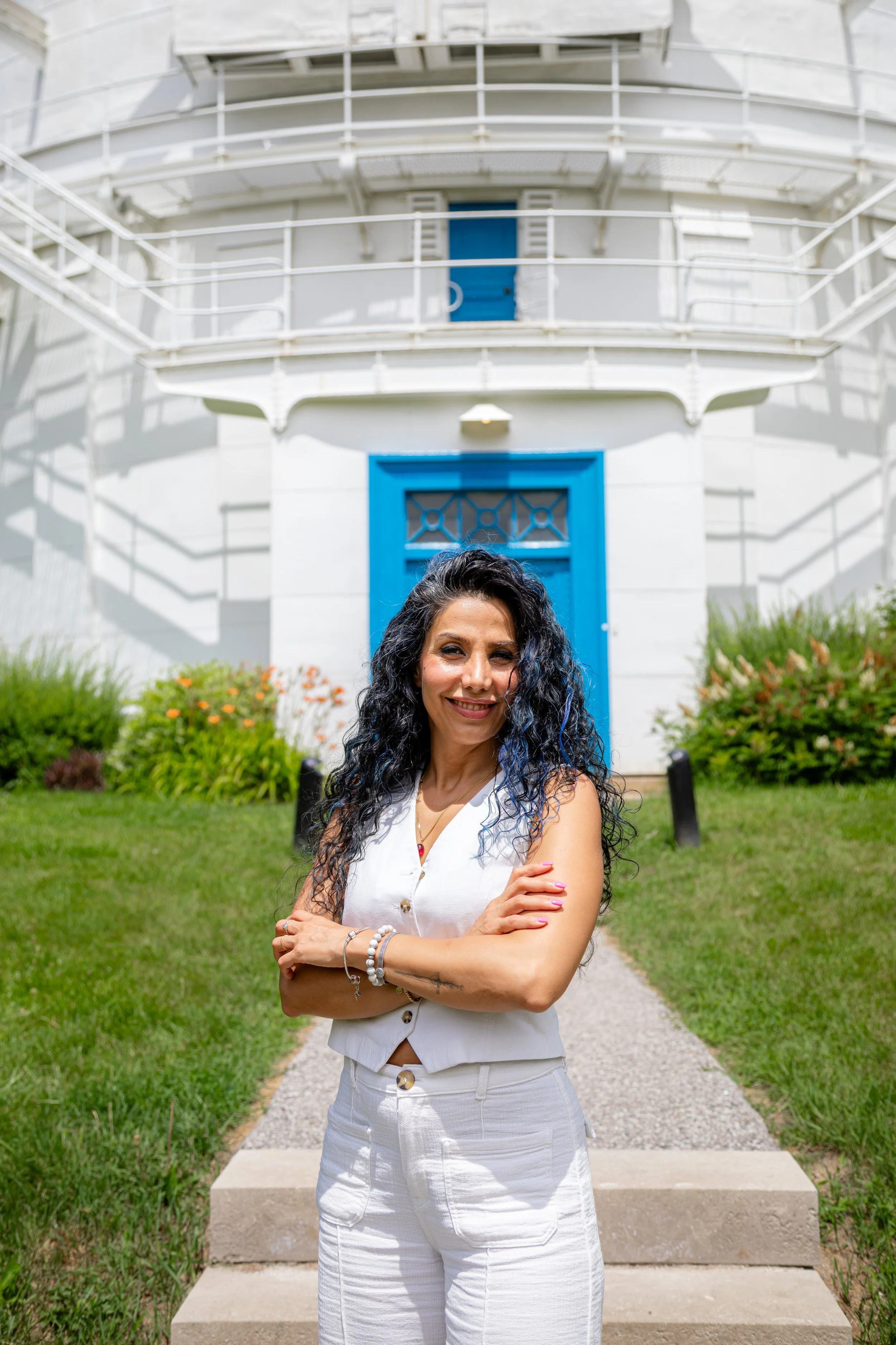A woman with curly black hair, wearing a white sleeveless top and white pants, stands outside on a gravel pathway with greenery and flowers on both sides. She is smiling with her arms crossed in front of her. Behind her is a building with a blue door, white exterior walls, and balconies with white railings.