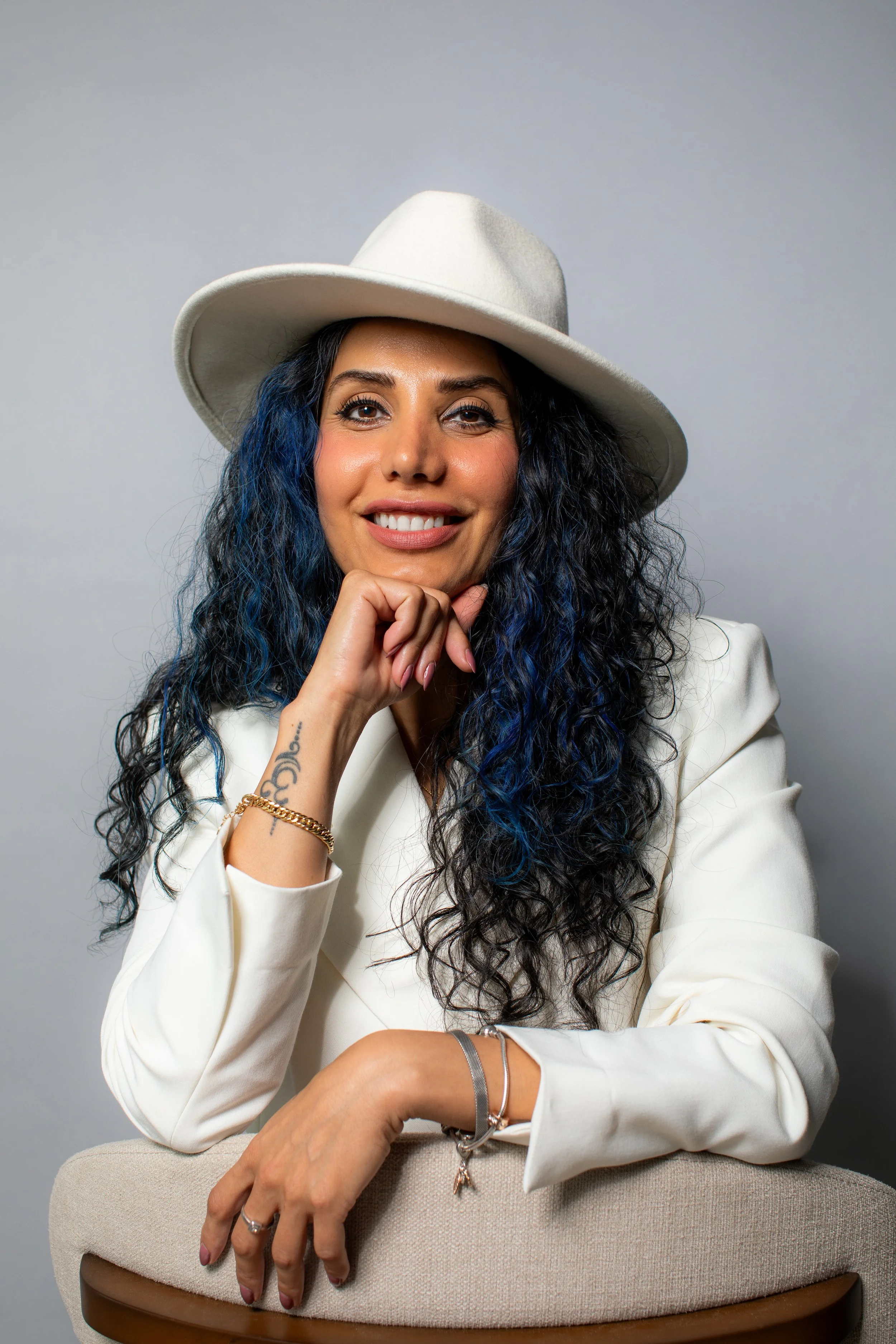 A woman with long, curly blue and black hair, wearing a white hat and white blazer, smiles and rests her chin on her hand, sitting on a beige stool against a plain gray background.