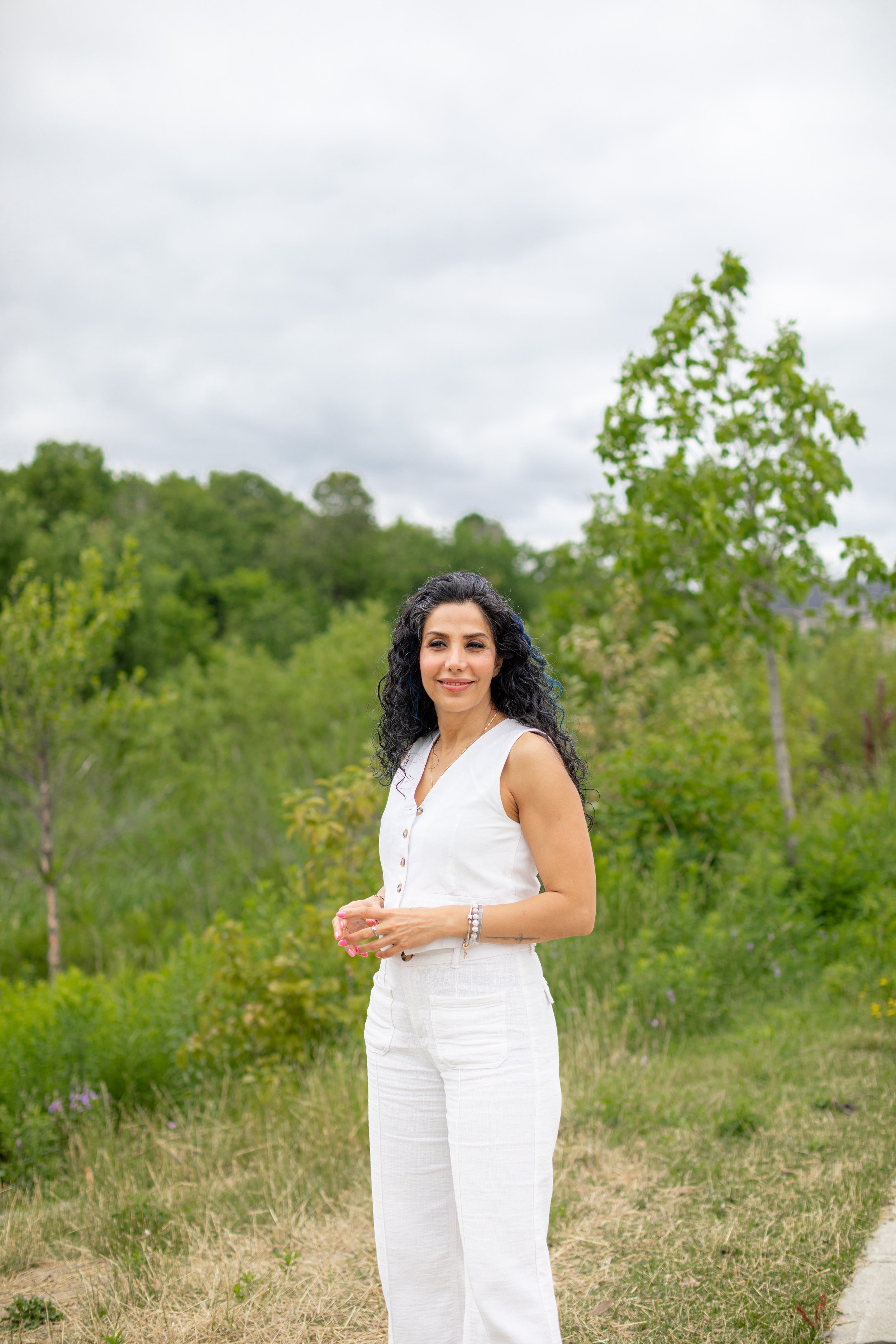 A woman with curly black hair, wearing a white sleeveless top and white pants, stands outdoors in a green park, smiling, with trees and cloudy sky in the background.