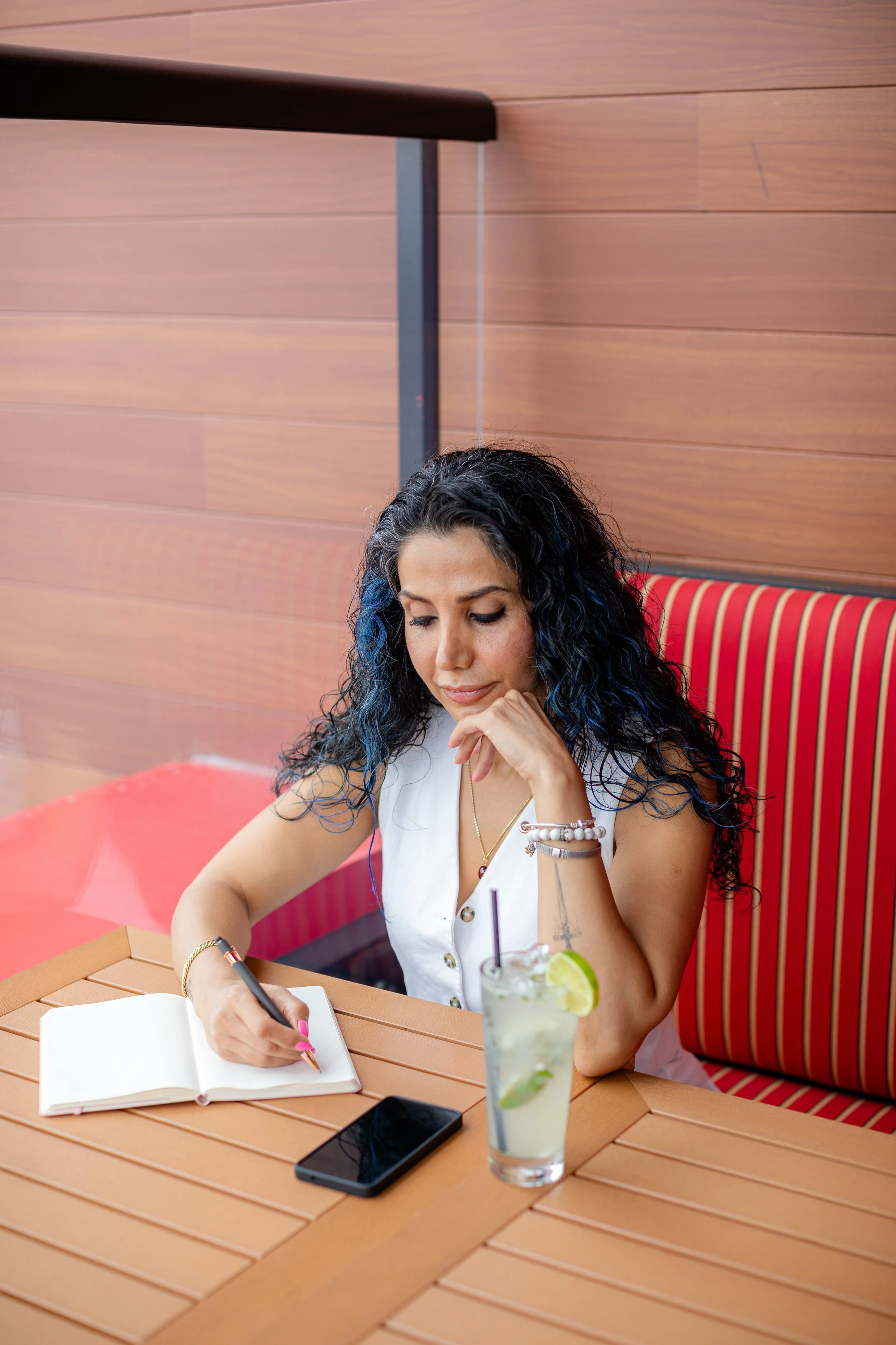 A woman with dark, curly hair with blue tips seated at a wooden table, writing in a notebook. She has a drink with lime and lemon slices, a straw, and ice in front of her. She is wearing a white sleeveless top and jewelry, and appears to be deep in thought.