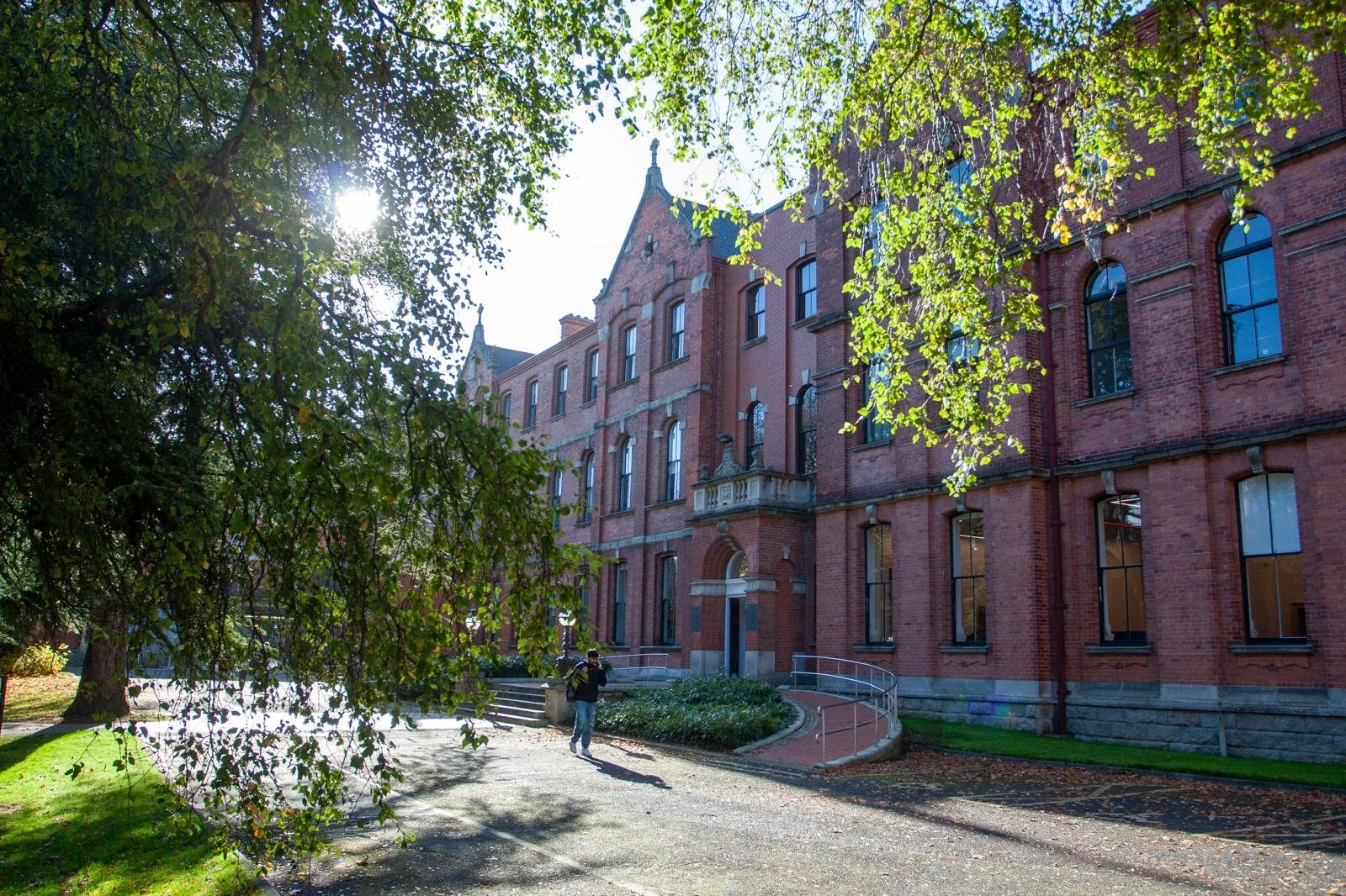 Autumnal view of the UCD Michael Smurfit Graduate Business School's red-brick Victorian building framed by tree branches, with a student walking along the path