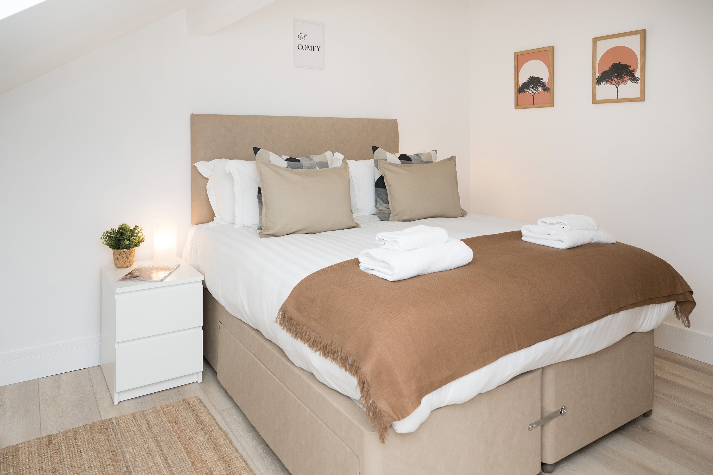 A neatly made bed with a beige upholstered headboard, six pillows with beige, white, and black-patterned pillowcases, and a brown blanket. On the right side, there are two stacks of white towels. A small white nightstand with a small potted plant, a 