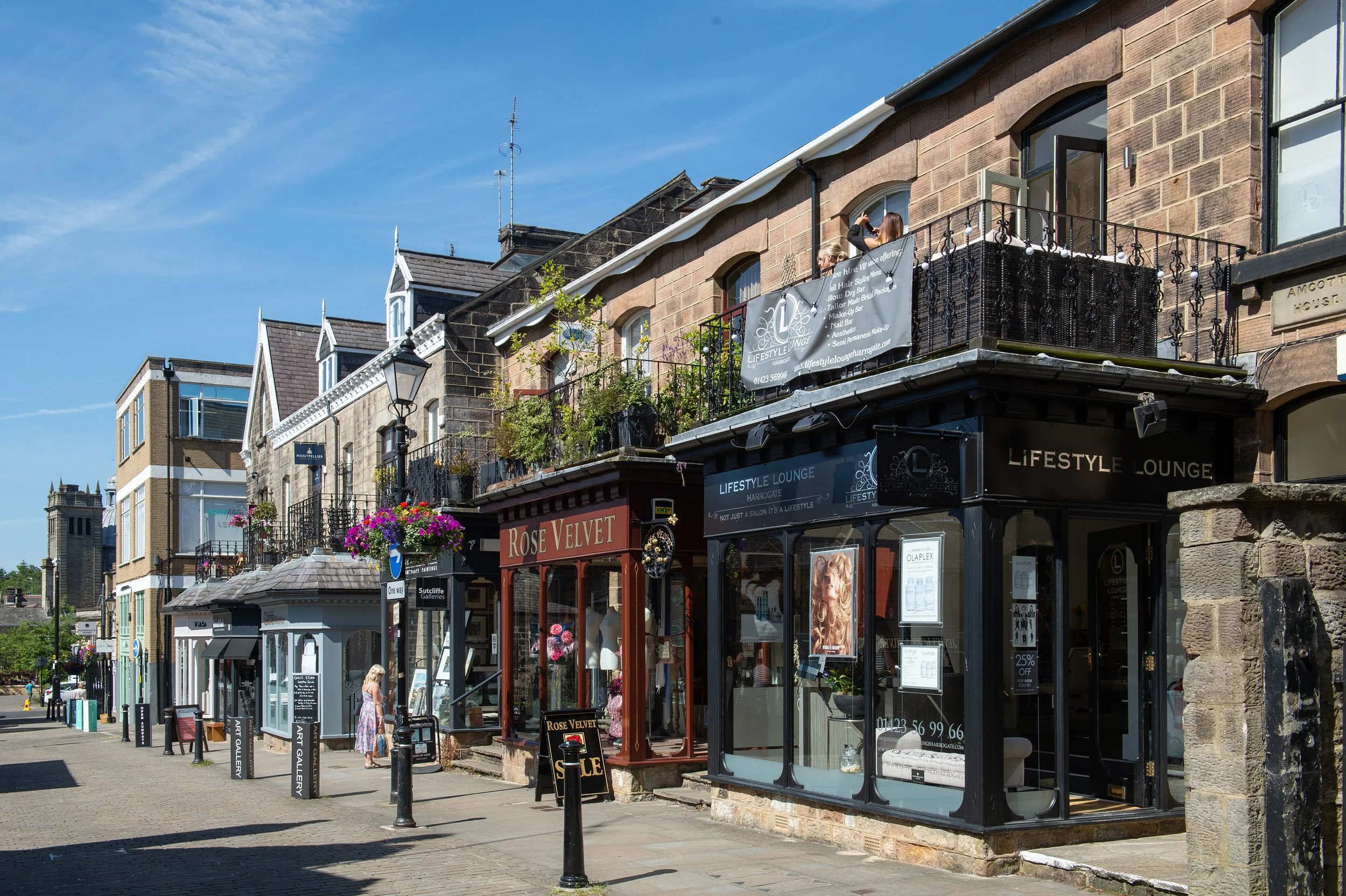 View of a street with shops, including a jewelry store, art gallery, and lifestyle lounge, with pedestrians walking and colorful flower baskets hanging outside. The buildings are multi-story with a mix of brick and stone facades under a clear blue sk