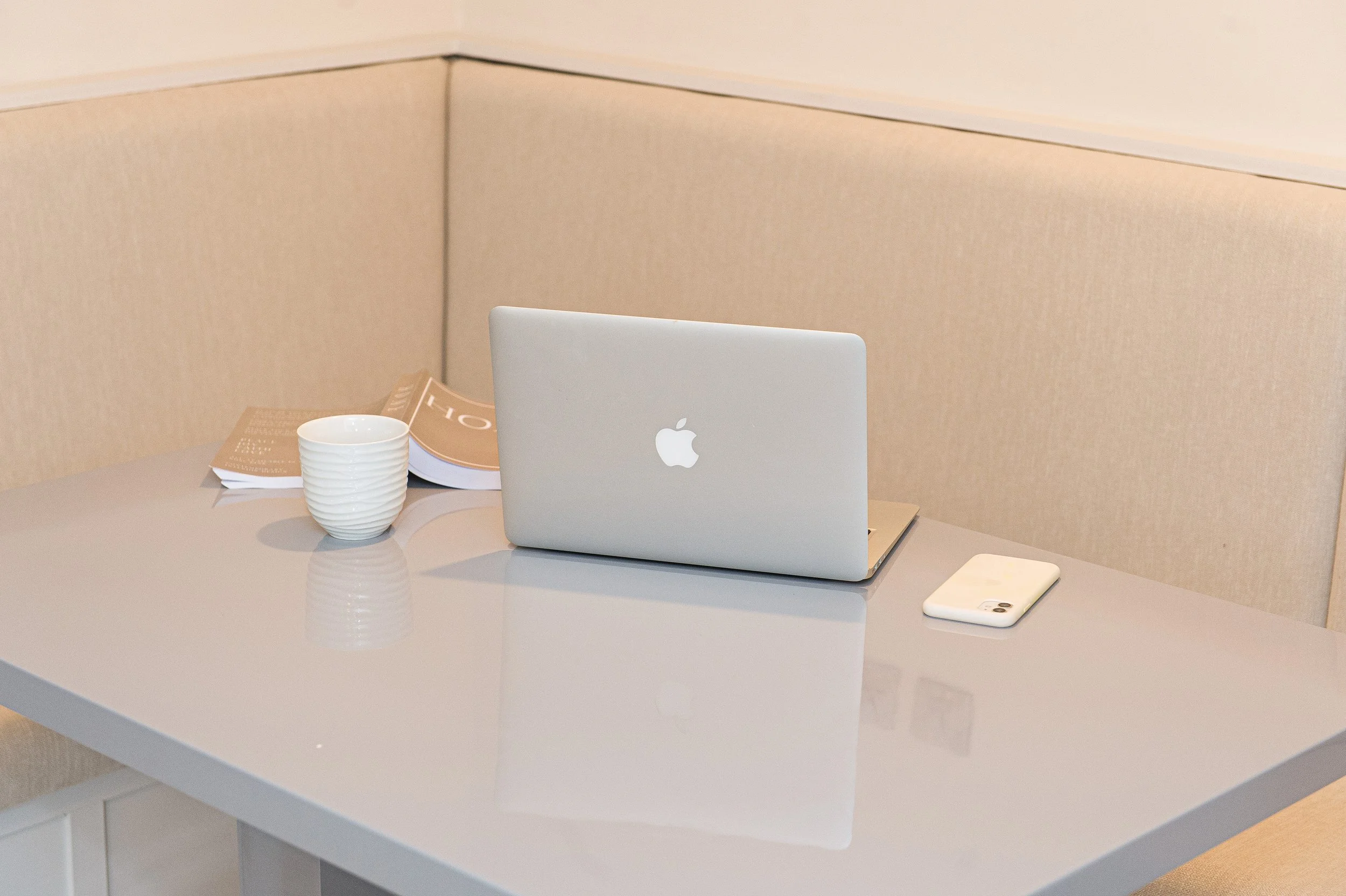 A minimalist workspace with a white Apple MacBook on a white table, a white smartphone, a white ribbed ceramic cup, and an open magazine, with a beige wall in the background.