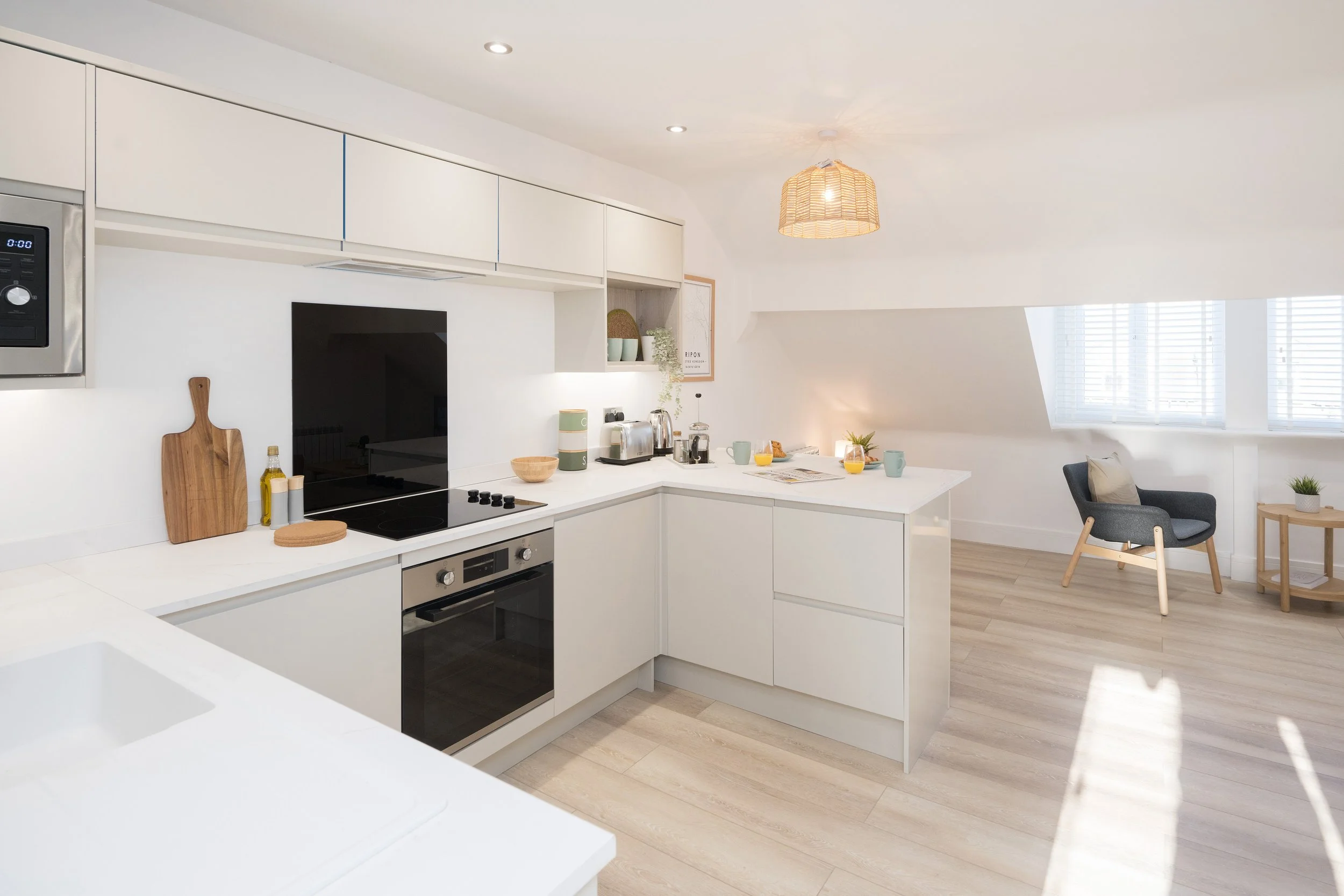 Modern white kitchen with built-in appliances, a dining table set for breakfast, a gray armchair, and large windows with blinds letting in natural light.