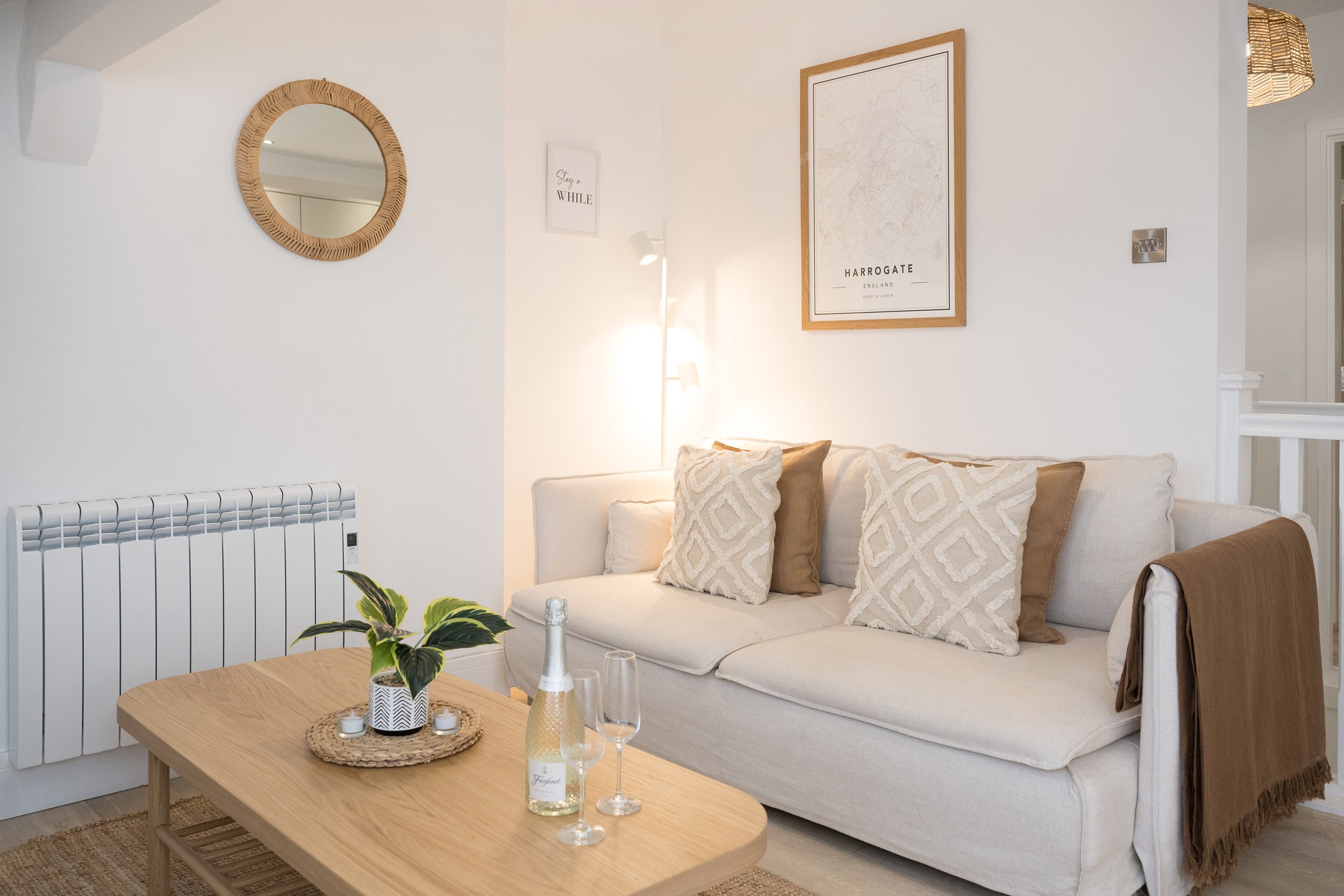 Living room with white walls, beige couch, patterned cushions, wooden coffee table with plant and candles, wall decor including a round wicker mirror, framed map of Harrogate, and a smaller framed quote, with soft lighting and a wicker lampshade.