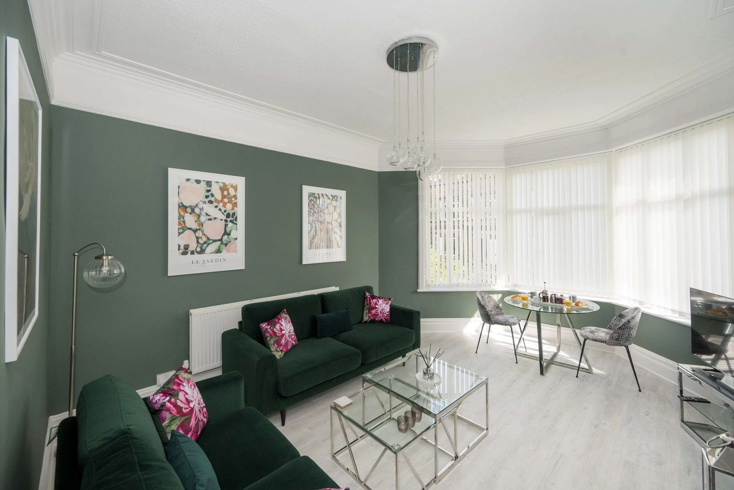 A modern living room with green walls, white ceiling and large bay window with vertical blinds, decorated with framed art and pink floral pillows, featuring a dark green sofa, glass coffee table, and a dining table with patterned chairs.
