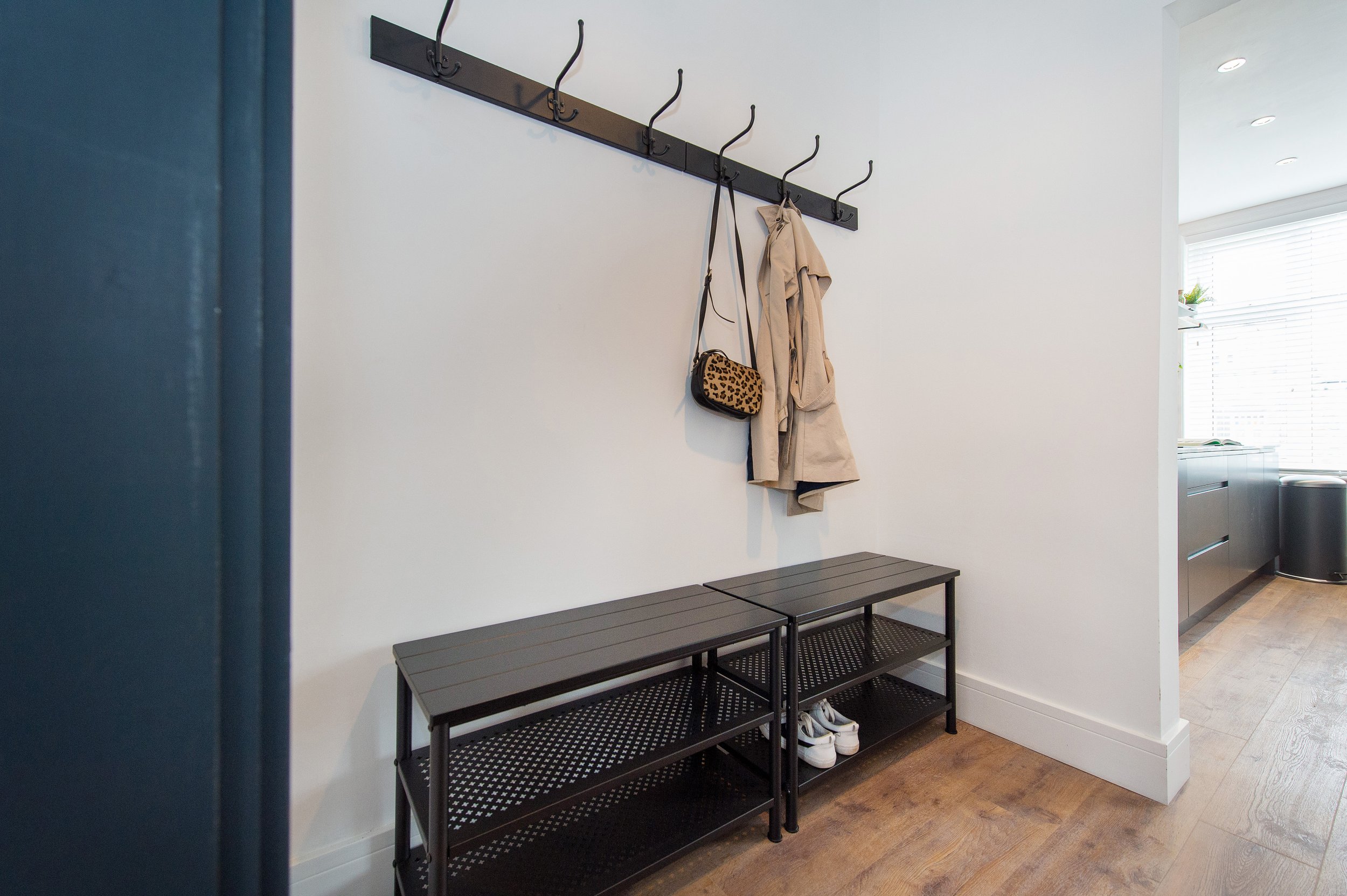 Entryway with black metal shoe rack, beige trench coat, and leopard print purse hanging on hooks on white wall, with black benches below and a window with blinds in the background.