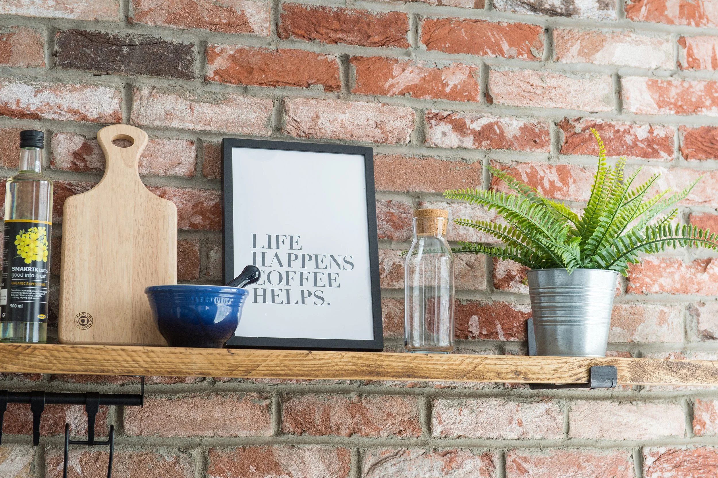 A wooden shelf mounted on a brick wall holding a clear glass bottle with a black cap, a wooden cutting board, an empty picture frame with text, a small glass jar, a blue bowl, and a potted fern in a metal container.