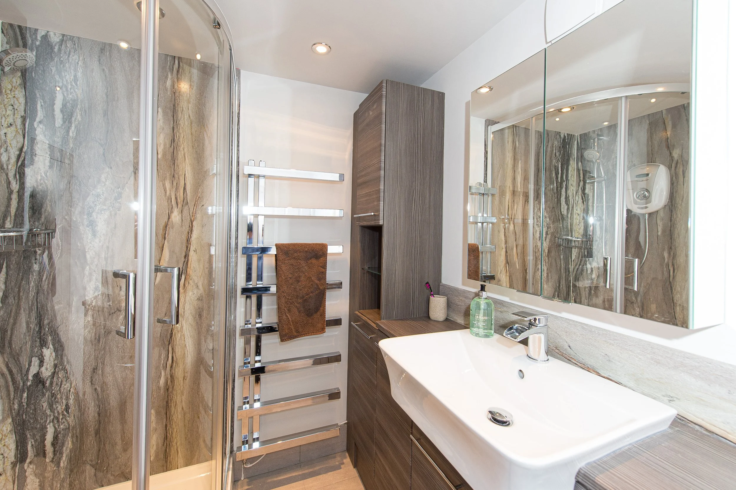 Modern bathroom with a glass shower enclosure, marble walls, a wooden cabinet, a large mirror, and a white countertop with a sink.