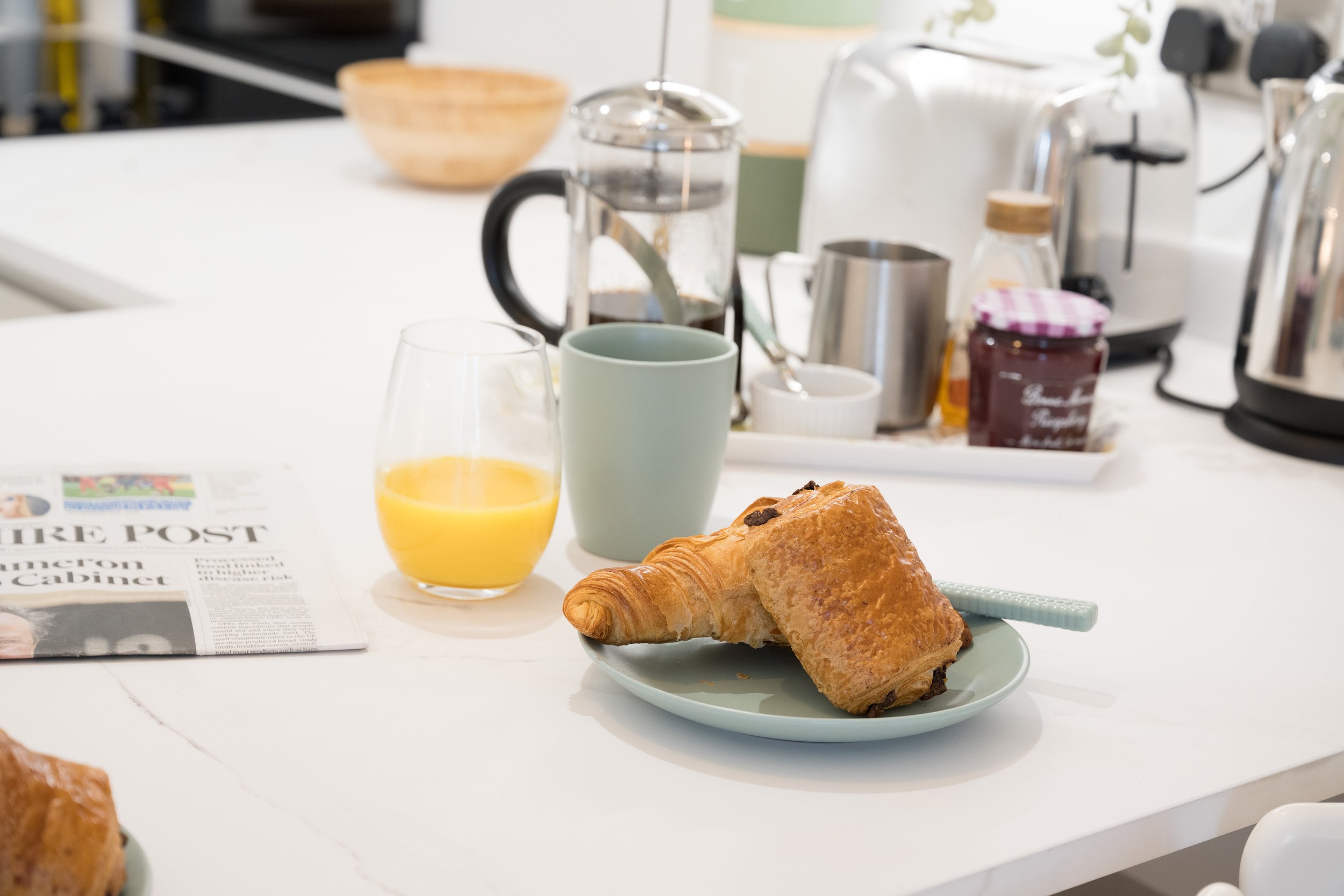 Breakfast setup on a white table with croissants on a plate, a glass of orange juice, a mug, and coffee-making appliances in the background.