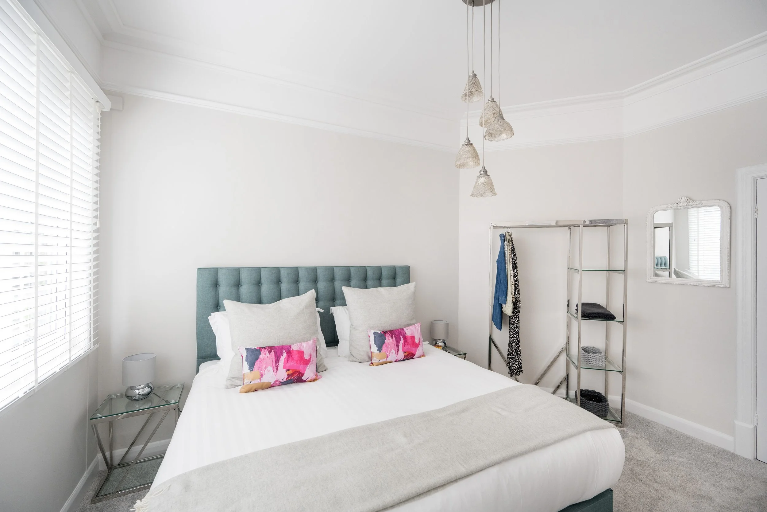 A bedroom with a white bed, colorful pillows, and a teal headboard. Two glass side tables with lamps, window with white blinds, a mirror, and a clothing rack with hanging clothes are visible.