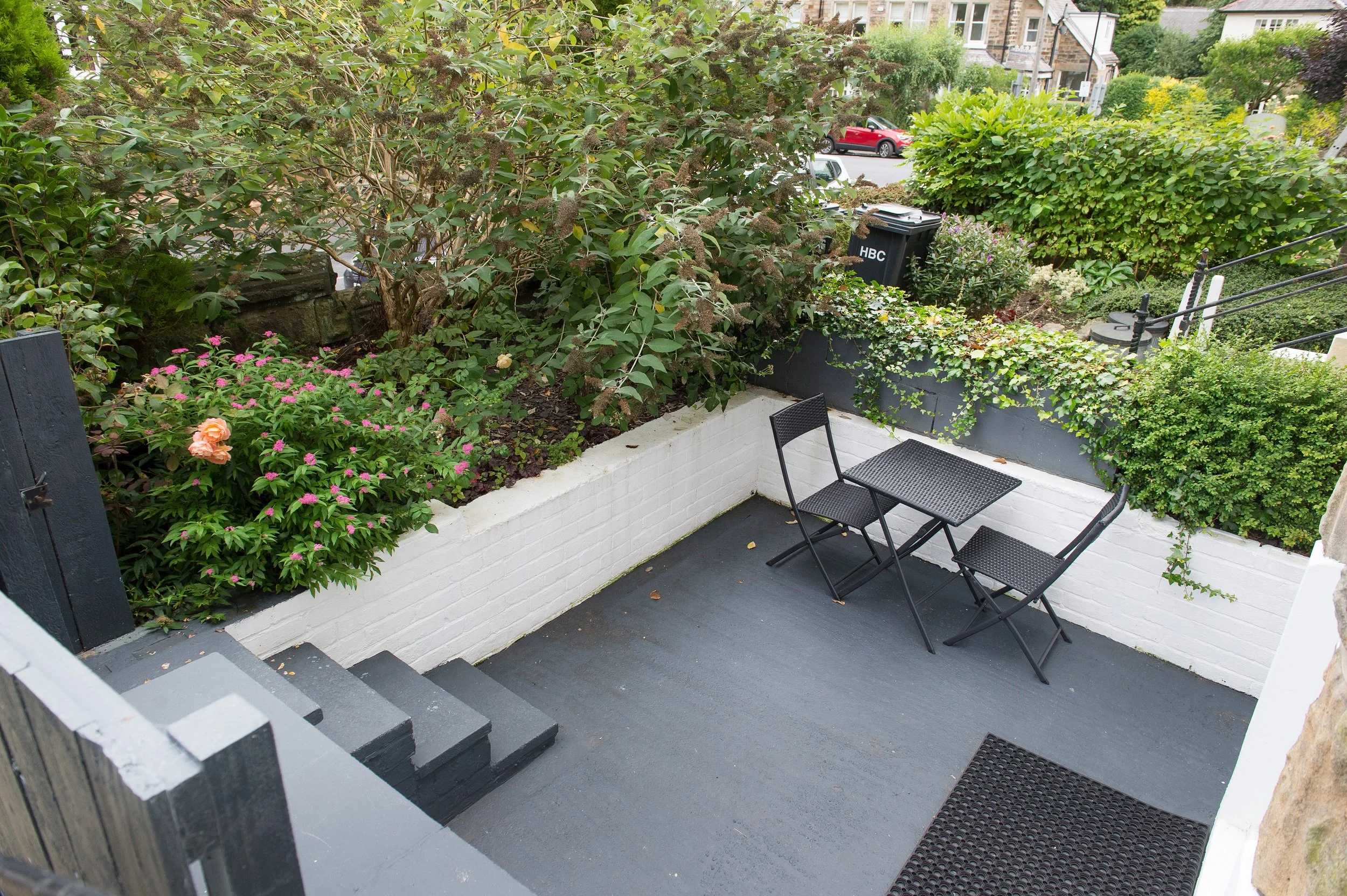 View of a small outdoor patio area with black metal chairs and plants, including bushes and flowers, enclosed by white brick walls and leading to a staircase.