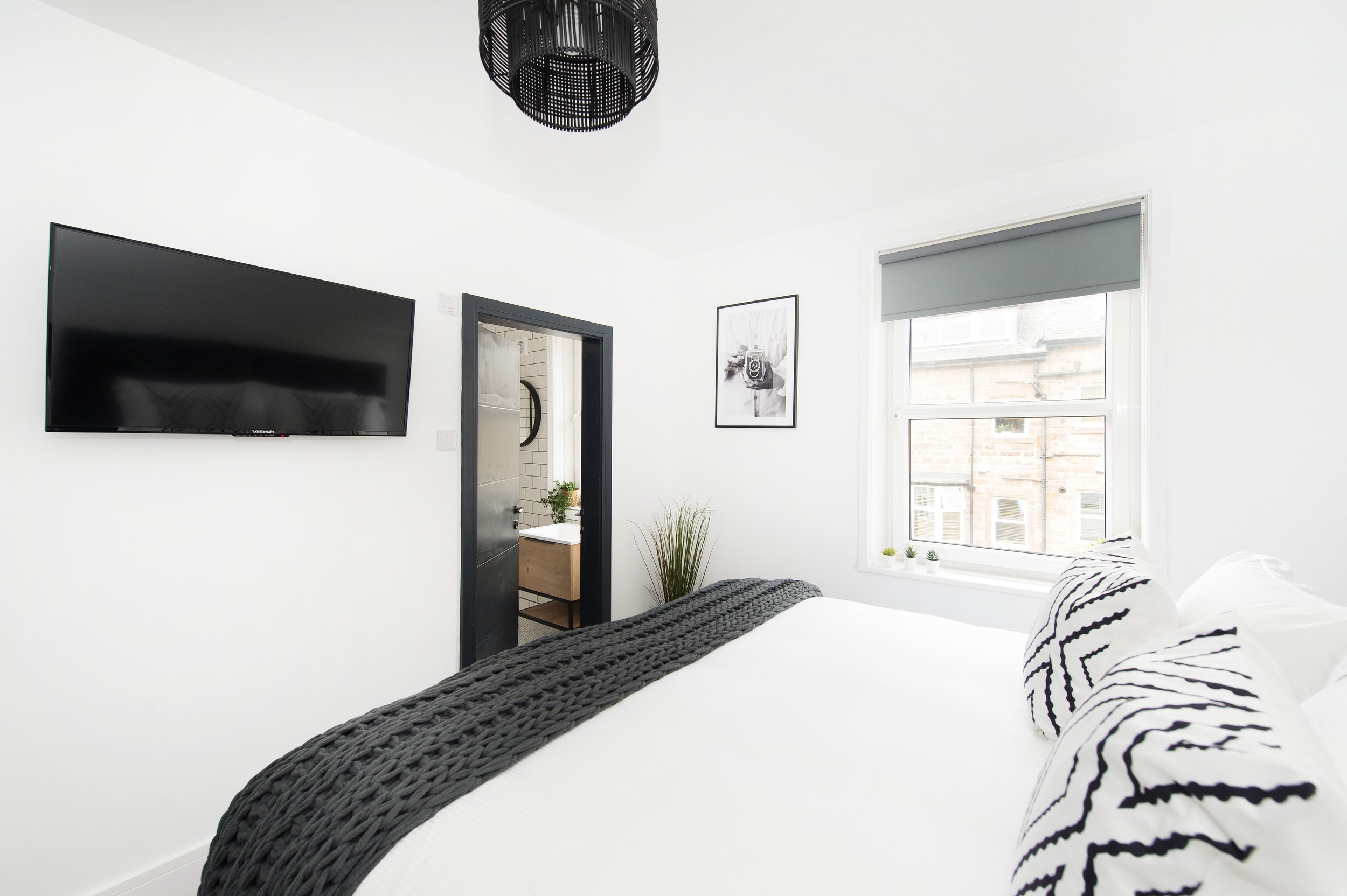 Modern bedroom with white walls, a wall-mounted flat-screen TV, a window with a gray blind, a framed black and white photograph, and a bed with black and white striped pillows.