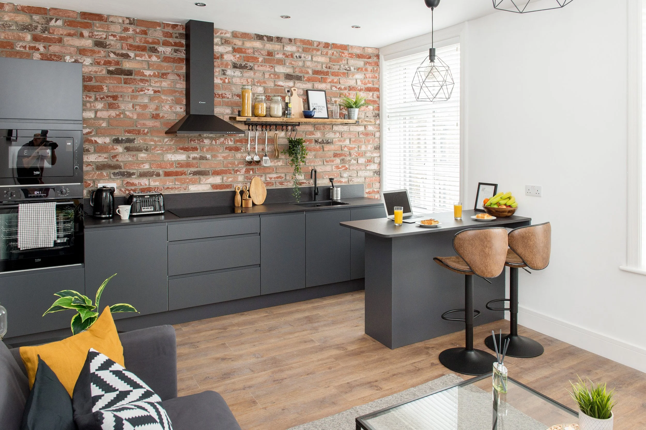 Modern kitchen with exposed brick wall, dark gray cabinets, and a small dining table with two chairs. Sunlight streams through window blinds, and there are breakfast items and a laptop on the table.