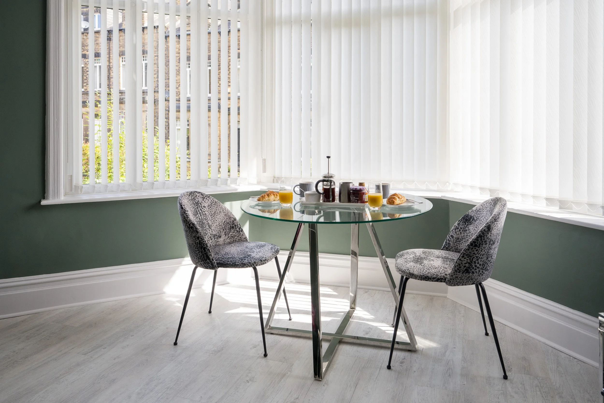 A round glass dining table set for breakfast with croissants, two glasses of orange juice, a French press, a mug, and condiments. Four leopard-print chairs surround the table inside a bright room with large windows and white vertical blinds.