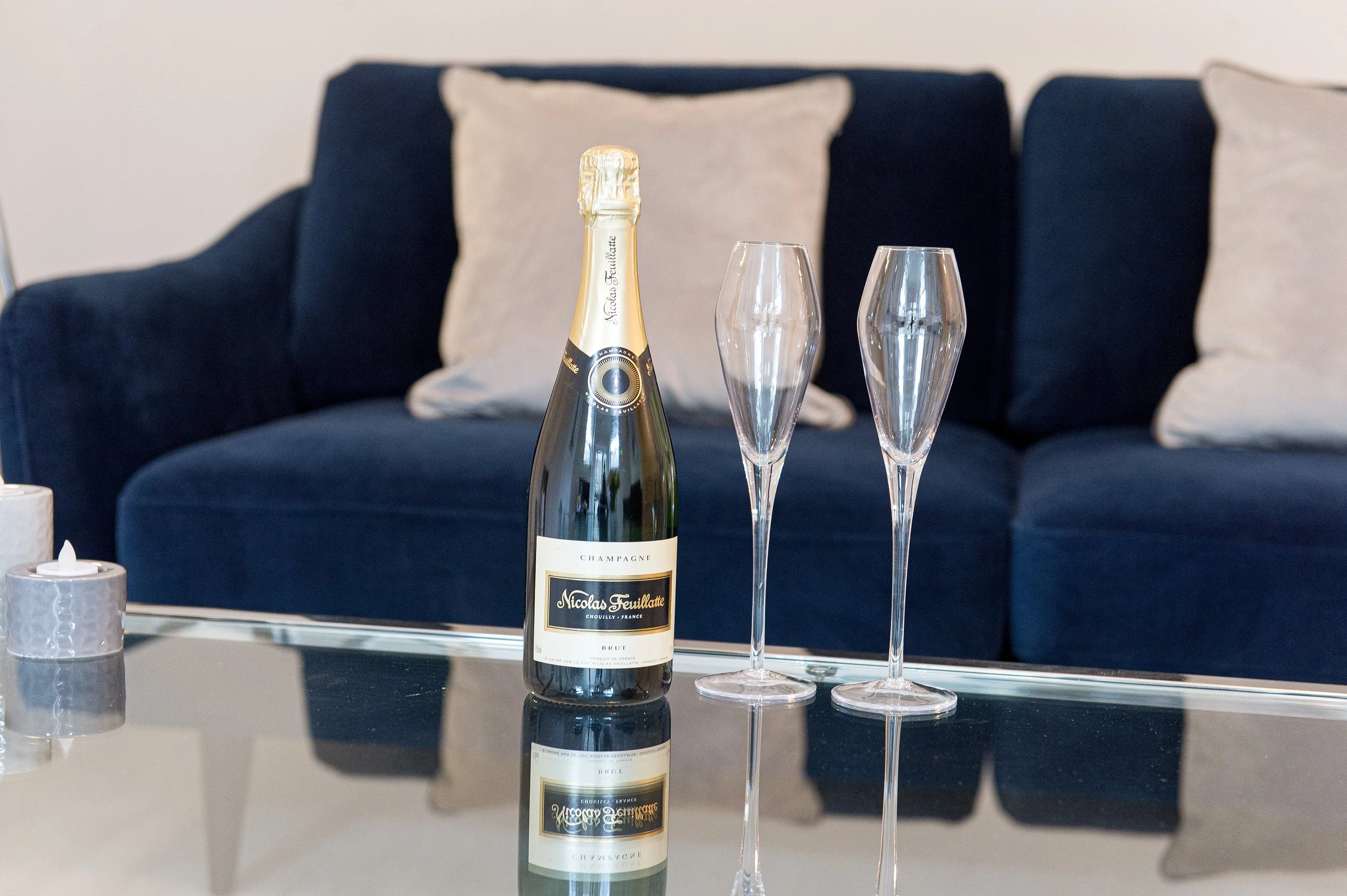 A bottle of Nicolas Feuillatte champagne and two empty champagne flutes on a reflective glass table in a living room with a blue sofa and white pillows.
