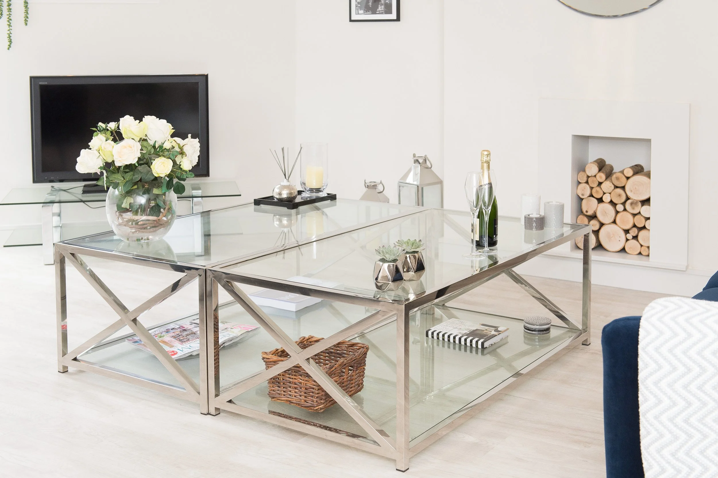 Modern living room with glass coffee table, floral arrangement, candles, bottles of sparkling water, and a TV, with a wood log storage nook in the background.