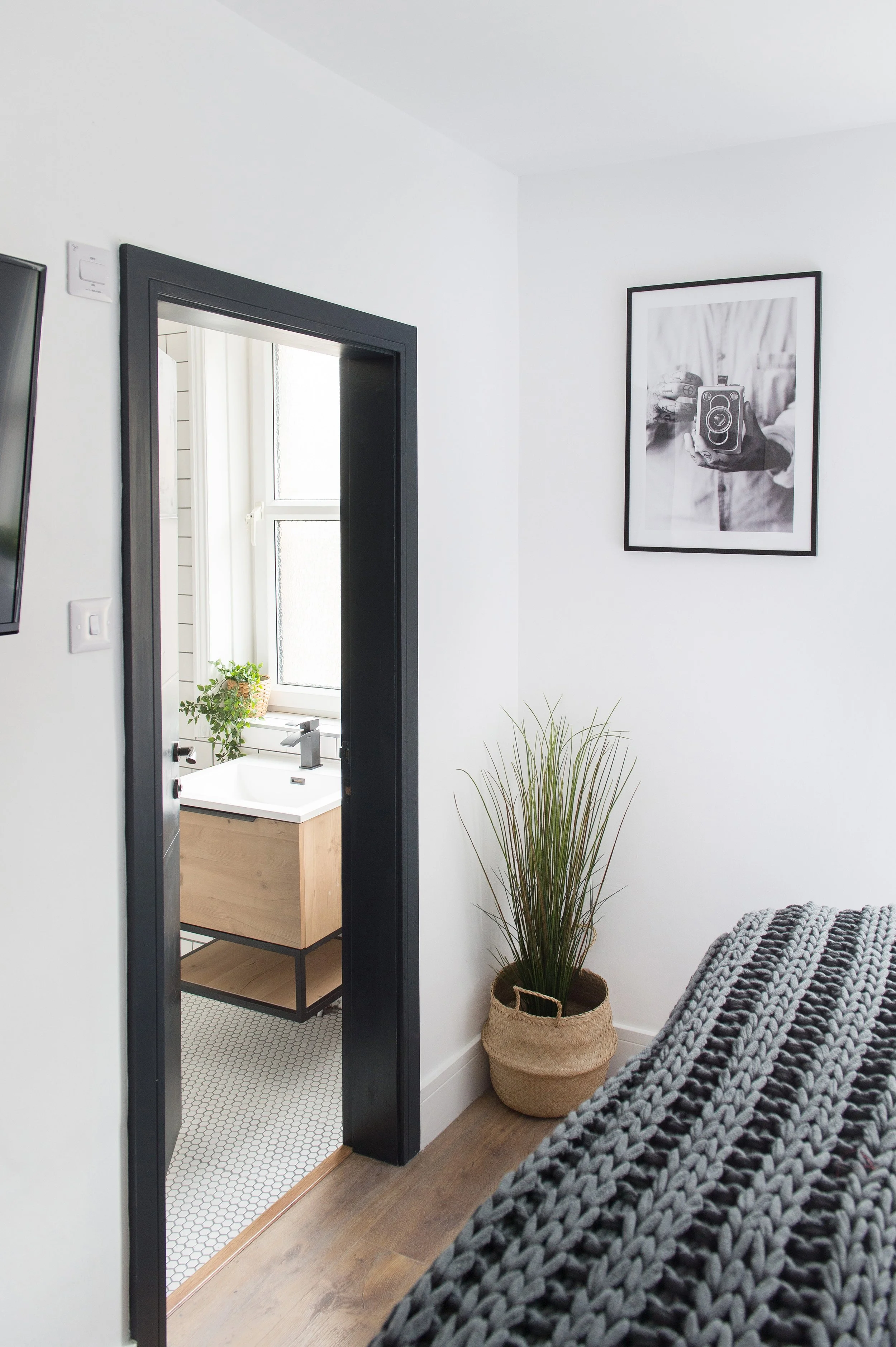 A bedroom with a bed covered in a chunky gray knit blanket, a large potted plant next to the bed, a black-framed framed photograph of a person holding a camera on the white wall, and a doorway leading to a bright bathroom with a small wooden cabinet,