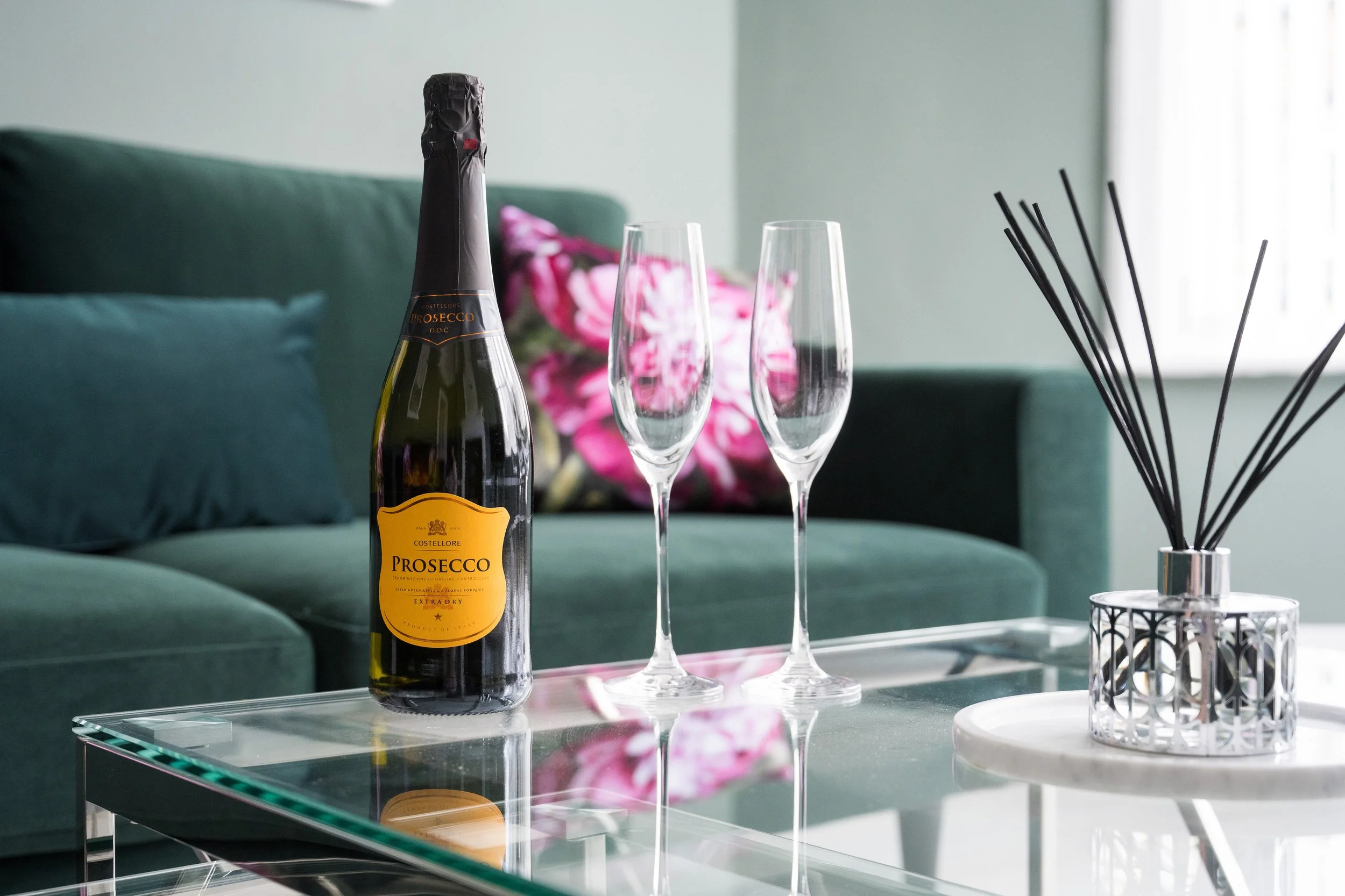 A bottle of Prosecco champagne and two empty champagne flutes on a glass table in a living room with green sofa and pink pillows.