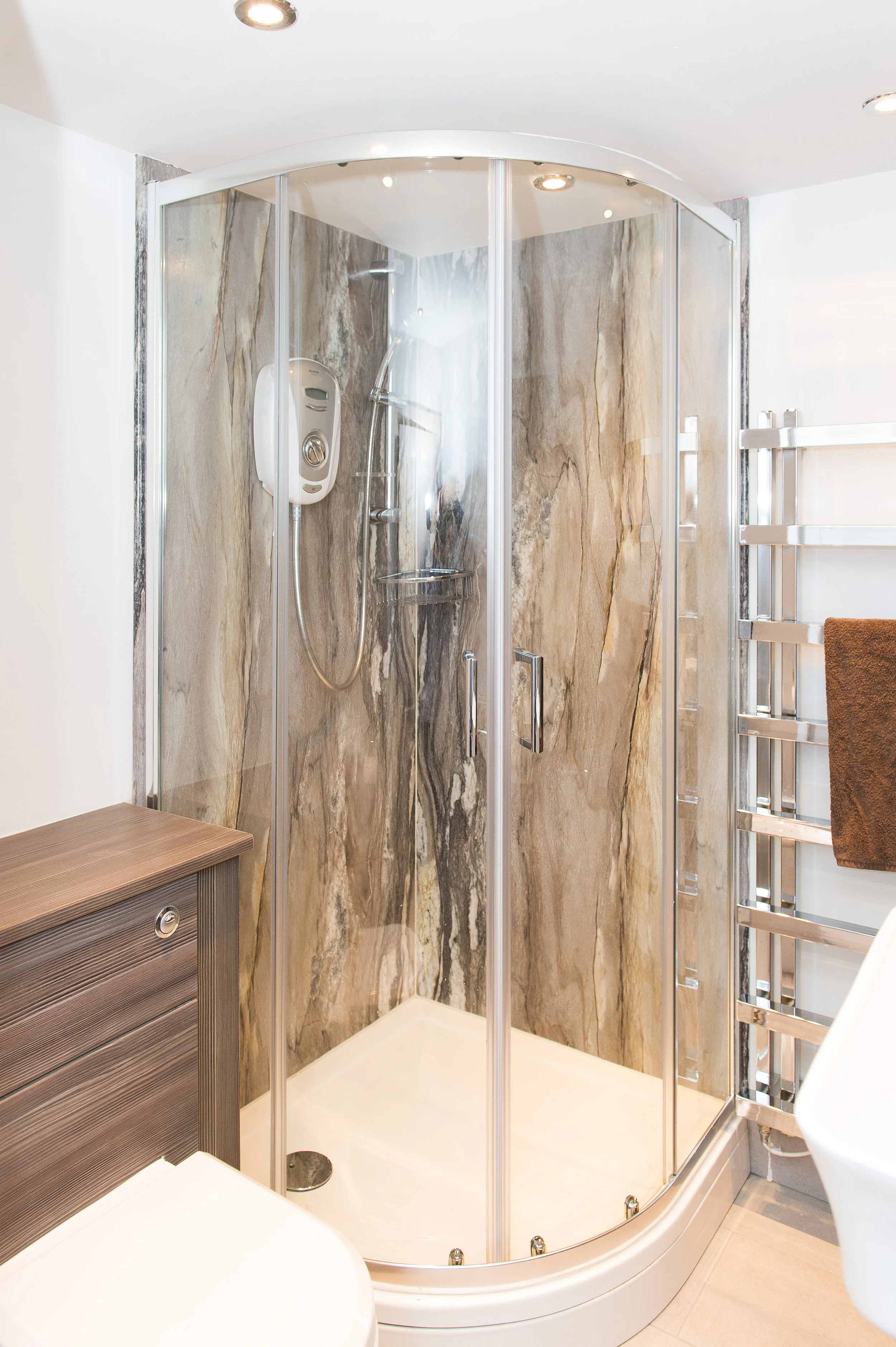 A modern corner shower with clear glass doors, marble wall tiles, a handheld showerhead, and a small shelf inside. Part of a white bathtub and wooden cabinet are visible nearby.