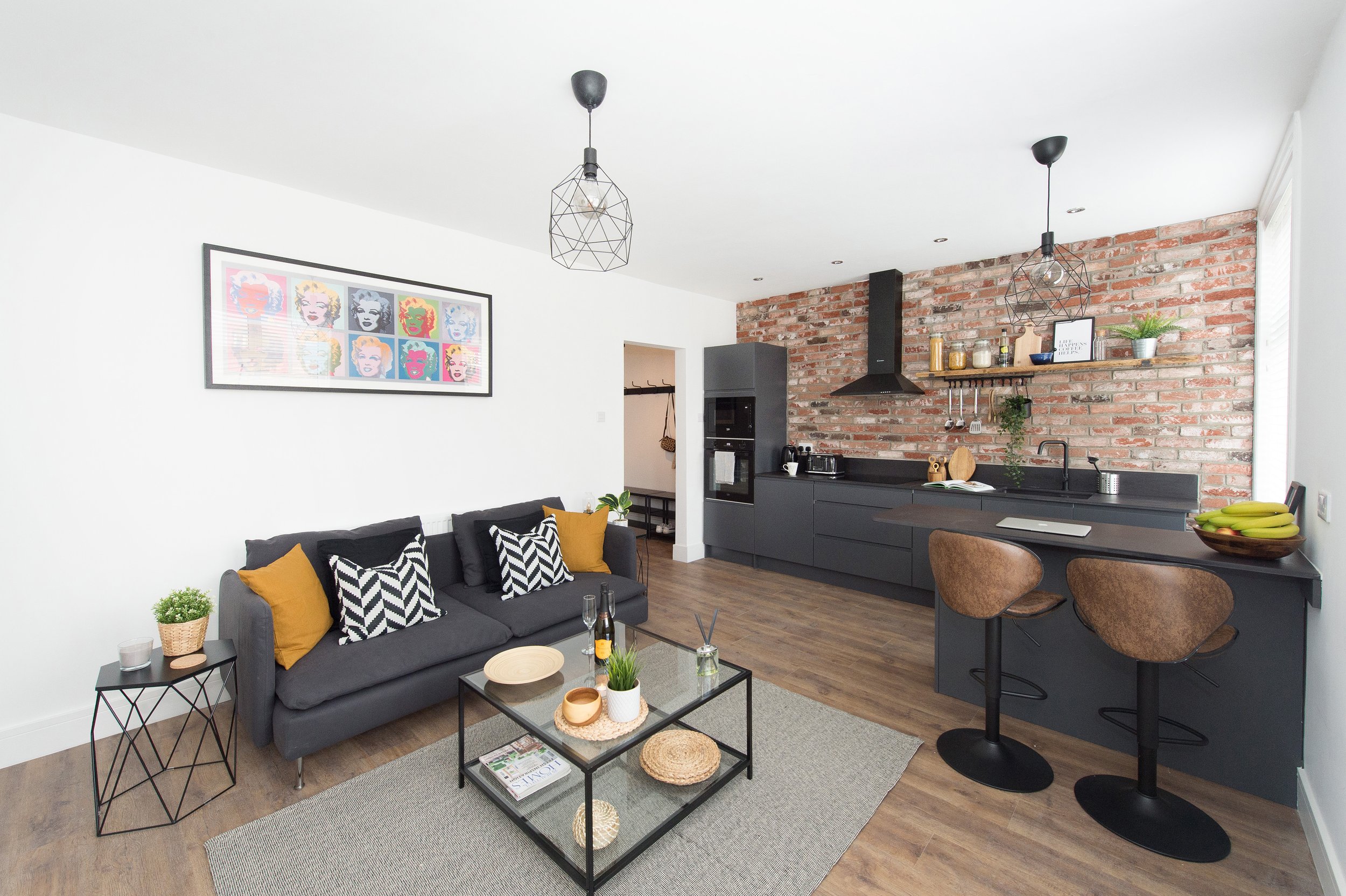 A modern open-concept living and kitchen area with minimalist decor. Features include a gray sofa with black and white patterned cushions, a black glass coffee table with decorative items, a brick accent wall, black cabinetry in the kitchen, and geom