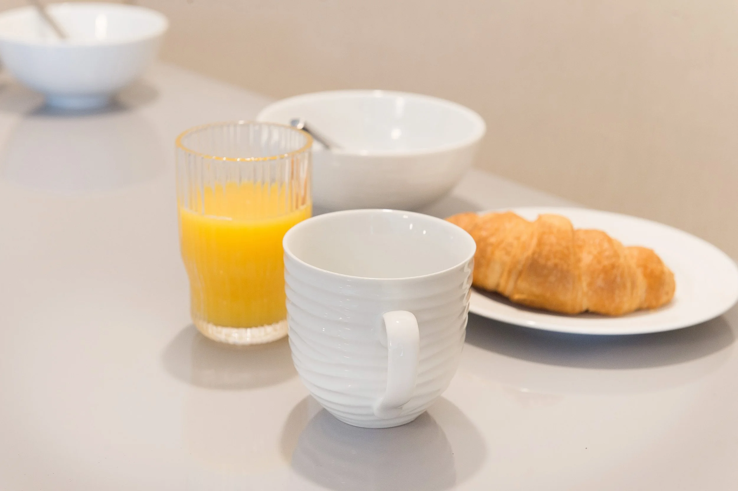 A breakfast table with a white mug, a glass of orange juice, a white bowl, and a plate with croissants.