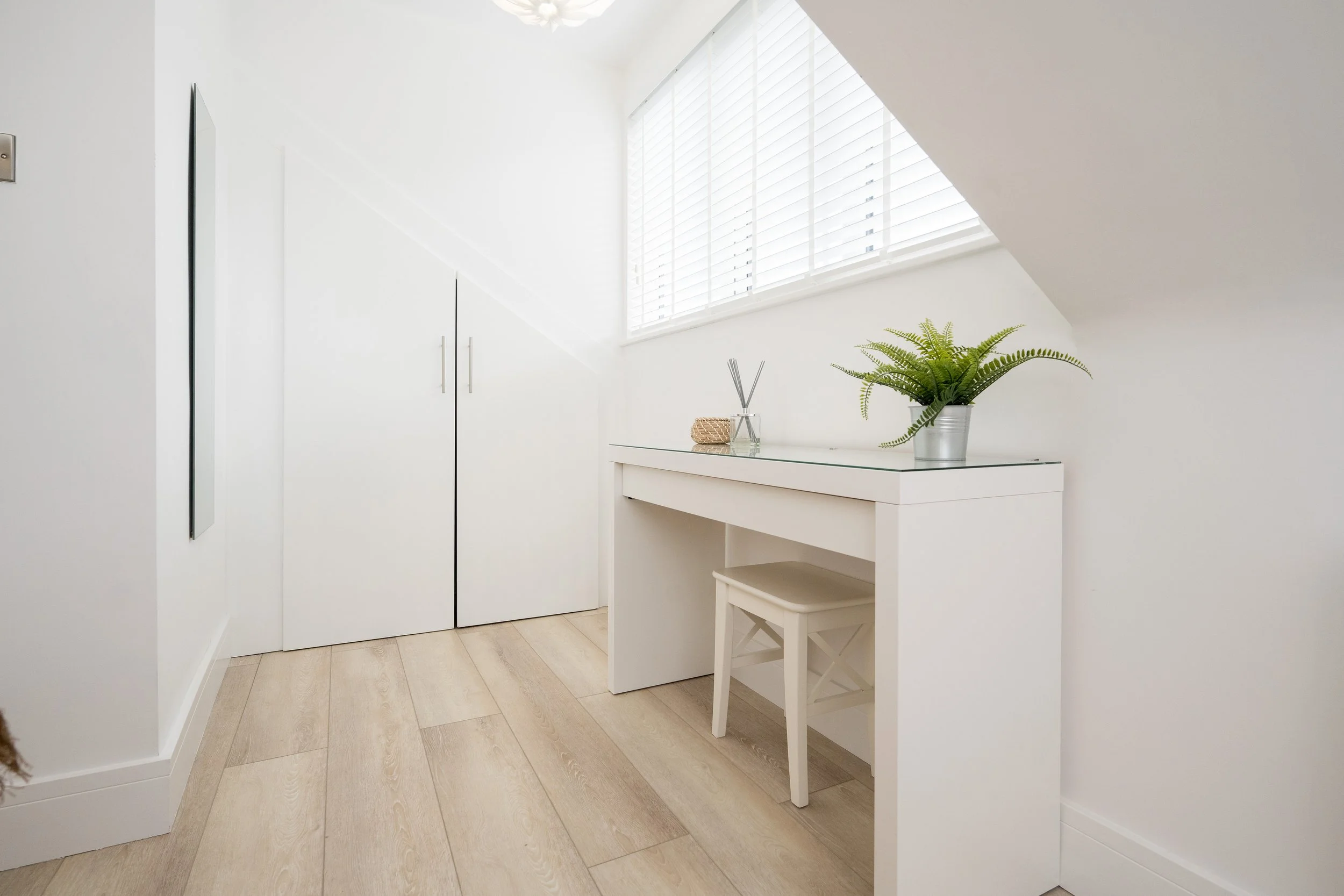 A minimalist white room with a large window with blinds, a white wardrobe, a small desk with a stool, a potted fern, and decorative items, with light wooden flooring.