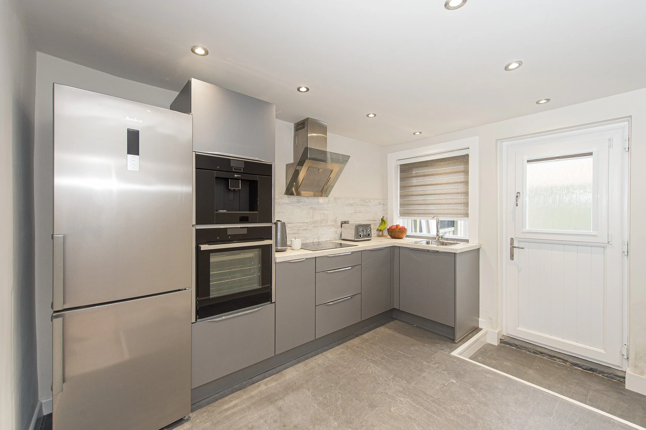 Modern kitchen with gray cabinets, stainless steel refrigerator, oven, and microwave, white marble countertops, a window with beige blinds, and a door, illuminated by ceiling recessed lights.