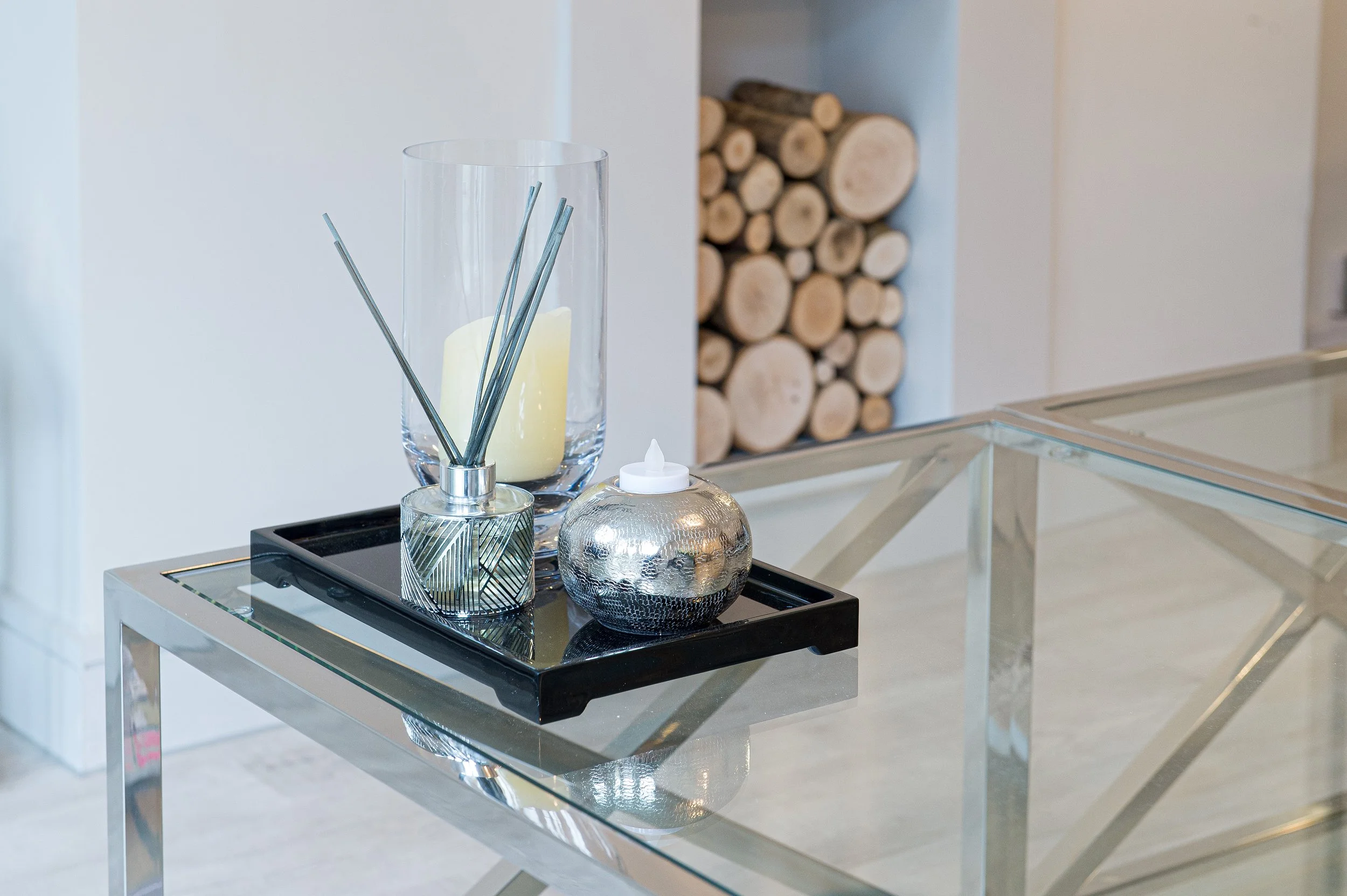 A glass table with decorative items, including a tall glass vase with a yellow candle and metal sticks, a small textured silver decorative object, and a small white bottle, with a background of a wall and a display of stacked firewood.