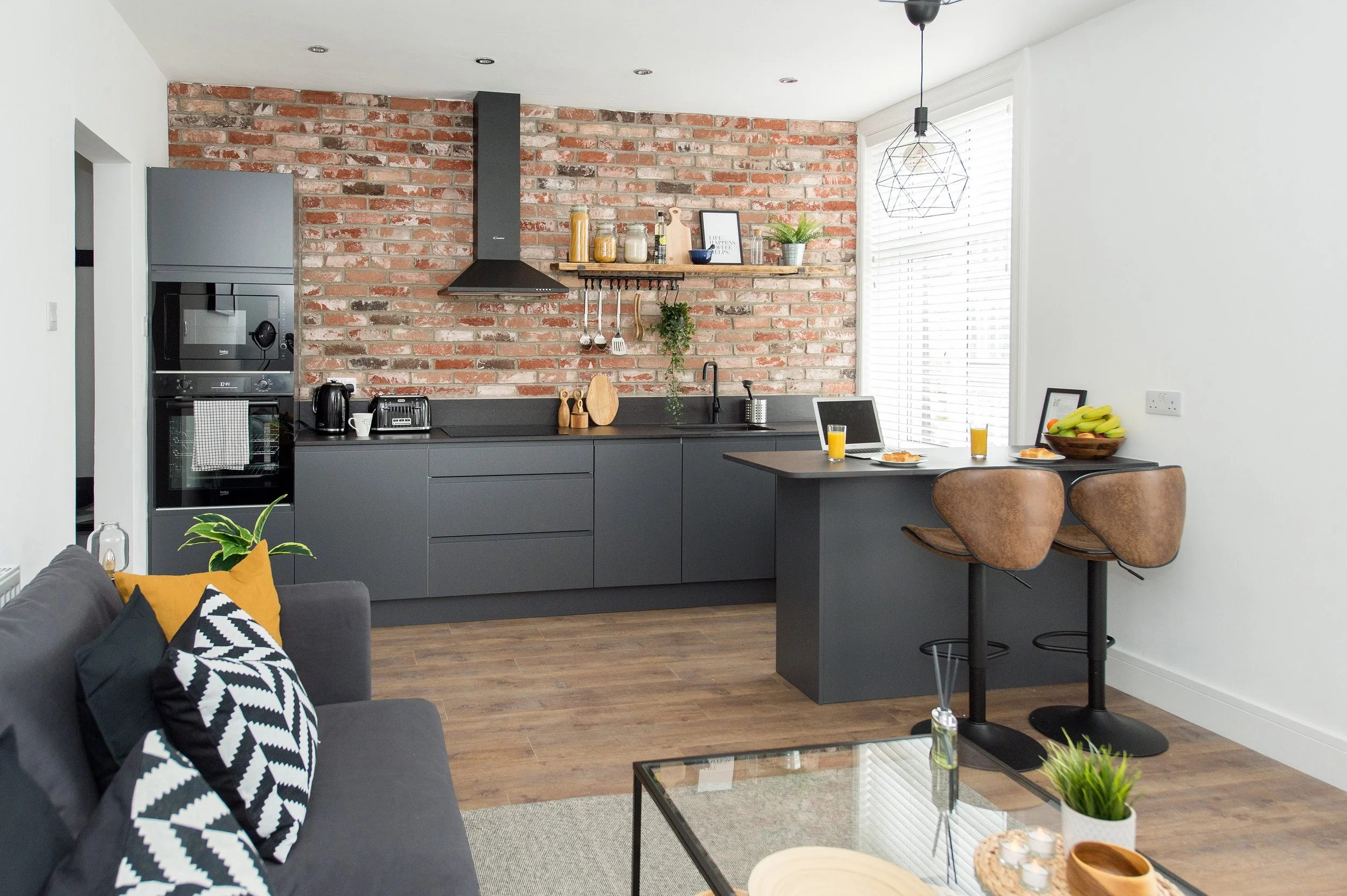 Modern kitchen with exposed brick wall, black cabinetry, and wooden accents. Countertop with kitchen utensils, a potted plant, and three glasses of orange juice. To the right, a dining area with two chairs and a fruit bowl. Part of a living space with a sofa and decorative pillows in the foreground.