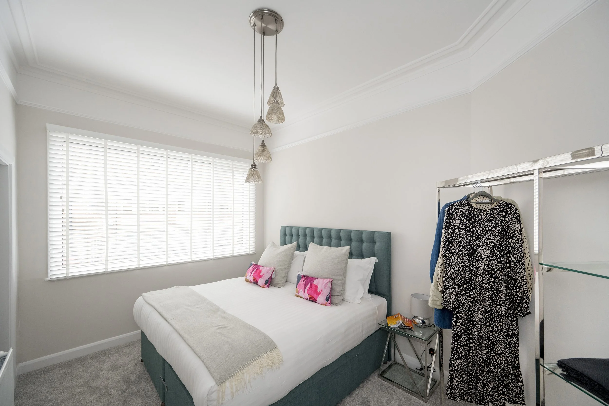 A bedroom with a bed, pillows, a window with blinds, a hanging light fixture, and a clothing rack with several garments.