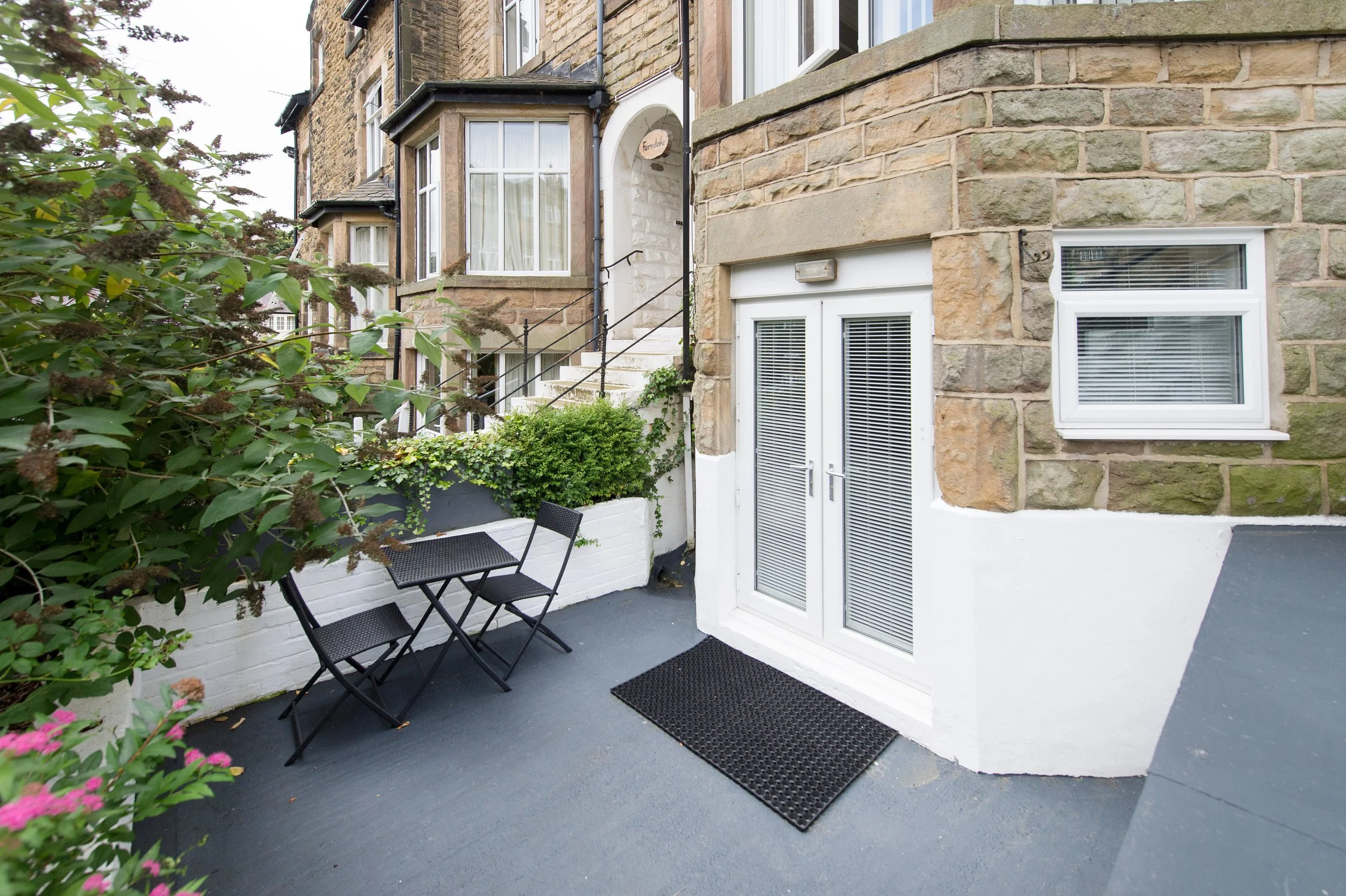 A small outdoor patio area with black foldable chairs, plants, and a wall with white-framed windows and a door, attached to a brick apartment building.