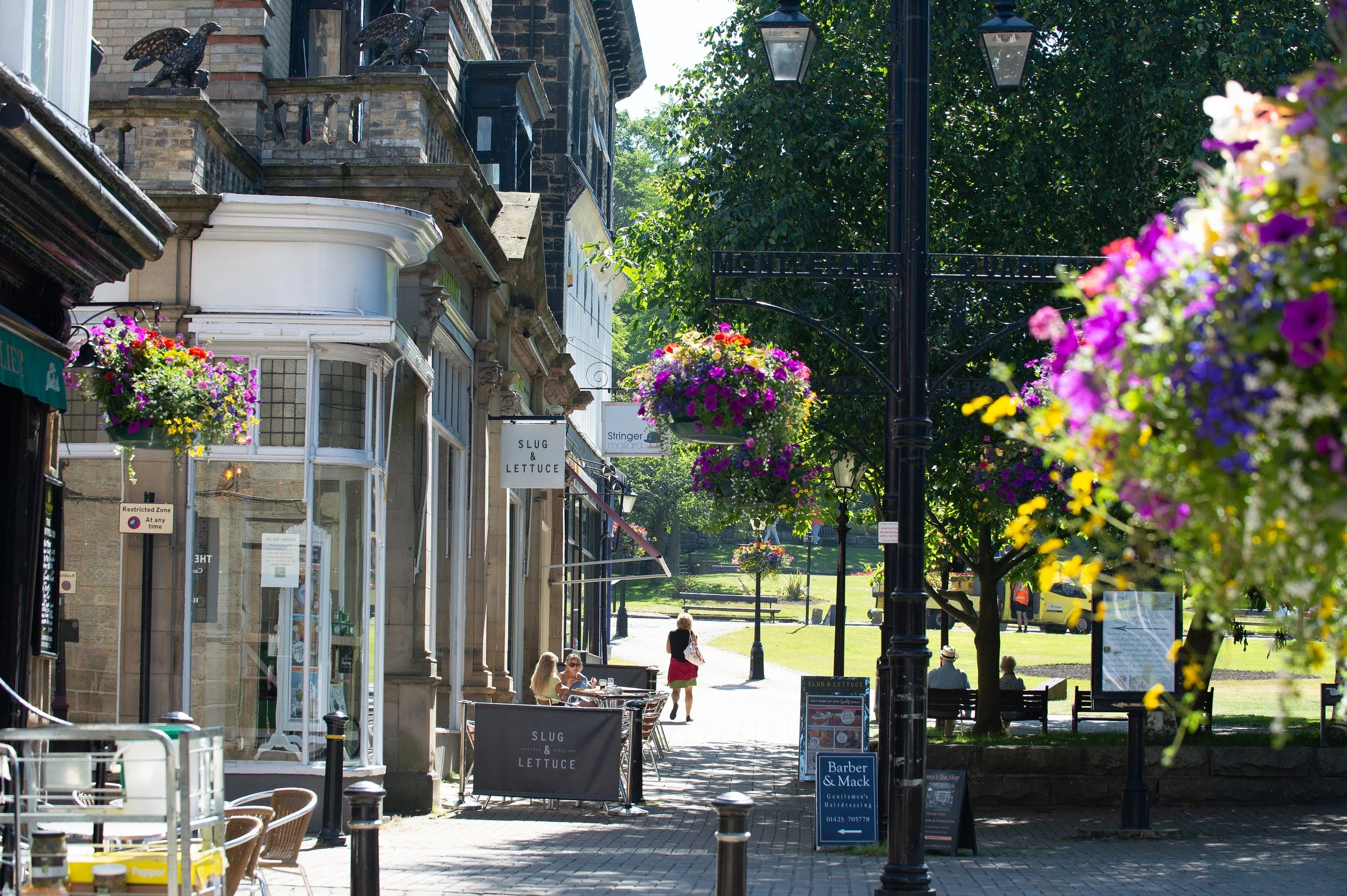 A lively street scene with shops, cafes, and colorful hanging flower baskets. People are walking and sitting outside, with trees and a park area in the background.