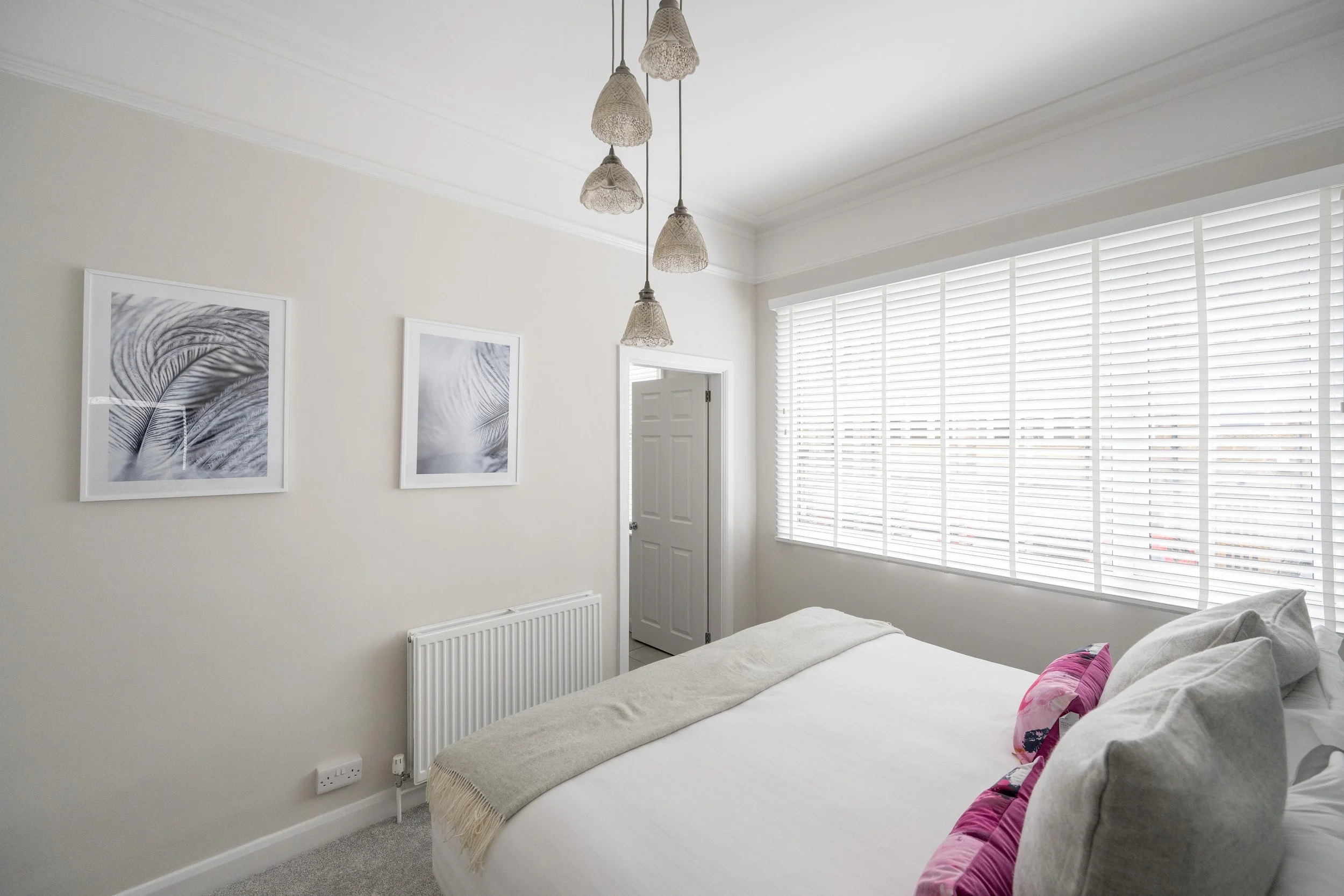 A bedroom with white walls, three hanging pendant lights with woven shades, and large white window blinds. There are two framed black and white leaf photographs on the wall, a white radiator below them, and a partially visible bed with white bedding 