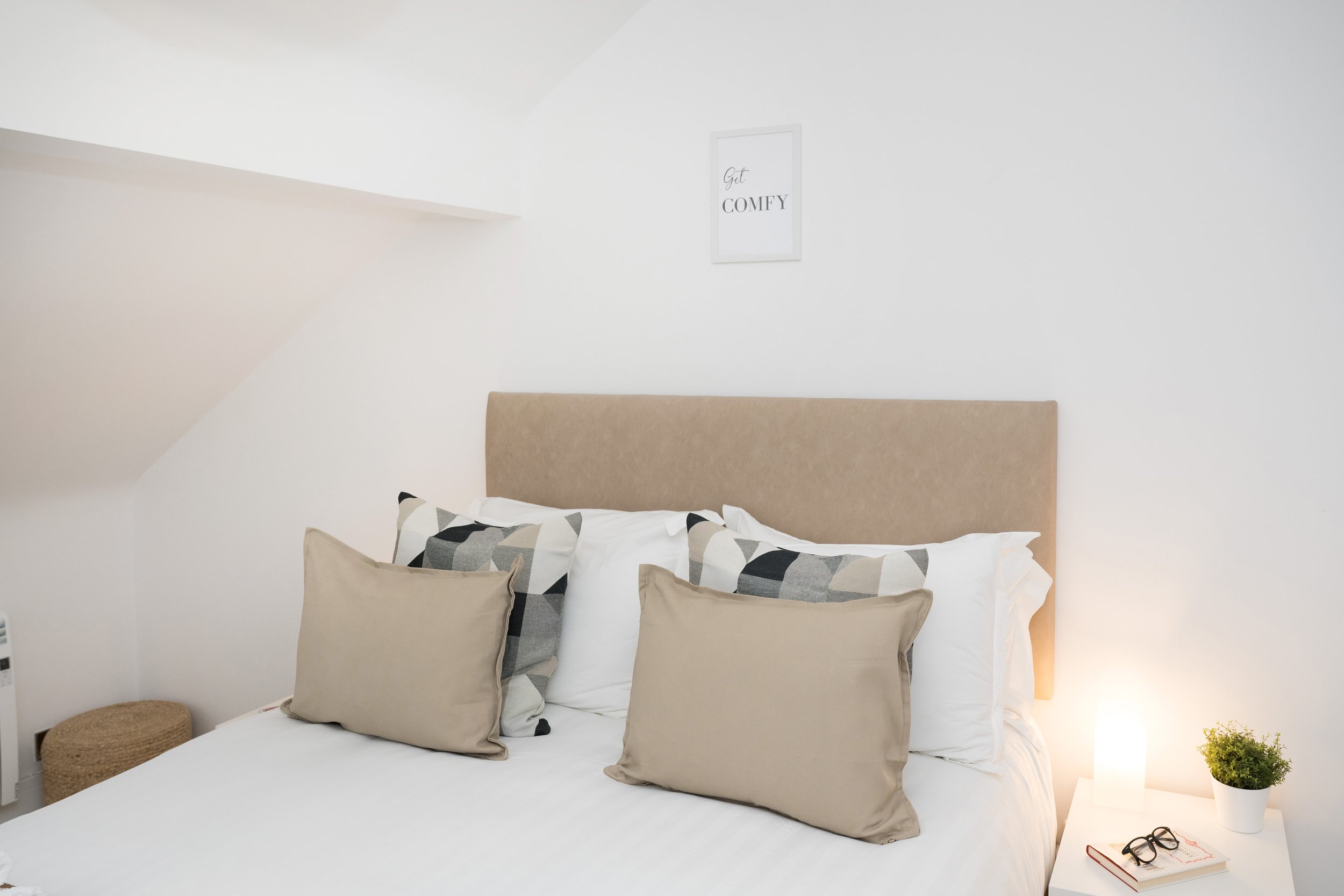 A neatly-made bed with a beige headboard, several pillows with beige, white, and black geometric patterns, a small beige cushion, and a bedside table with a potted green plant, a pair of glasses, and a notebook, in a bright, minimalistic room.