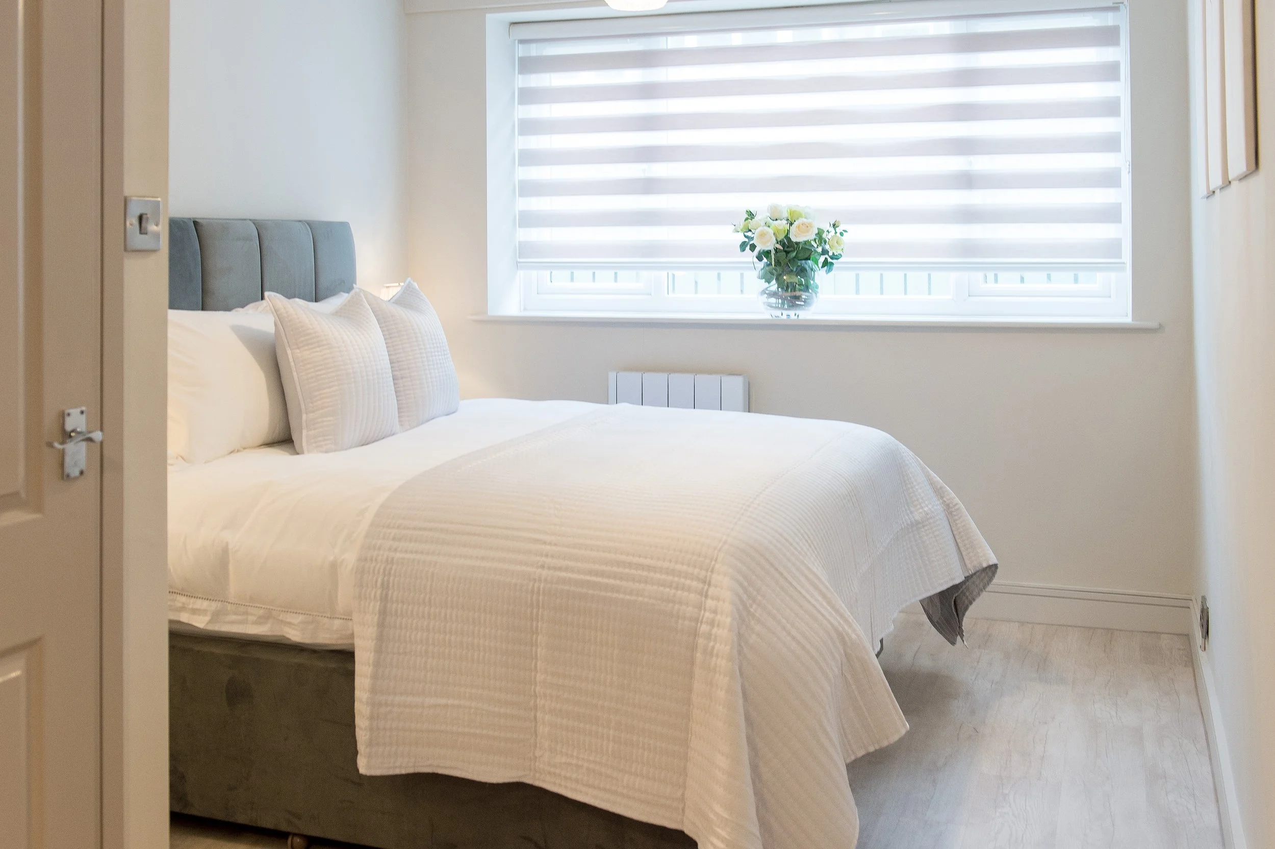 A neatly made bed with white bedding and matching pillows, a gray upholstered headboard, a window with horizontal blinds, and a small bouquet of white and yellow roses in a glass vase on the windowsill.
