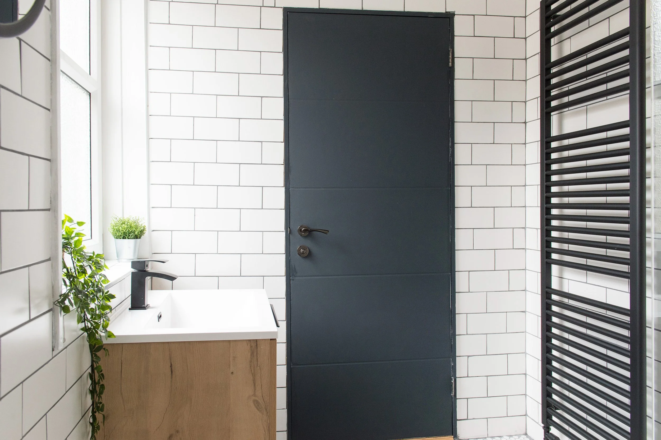 A modern bathroom with white subway tiles, a black door, a black towel rack, a white sink with a black faucet, and a small plant on the windowsill.