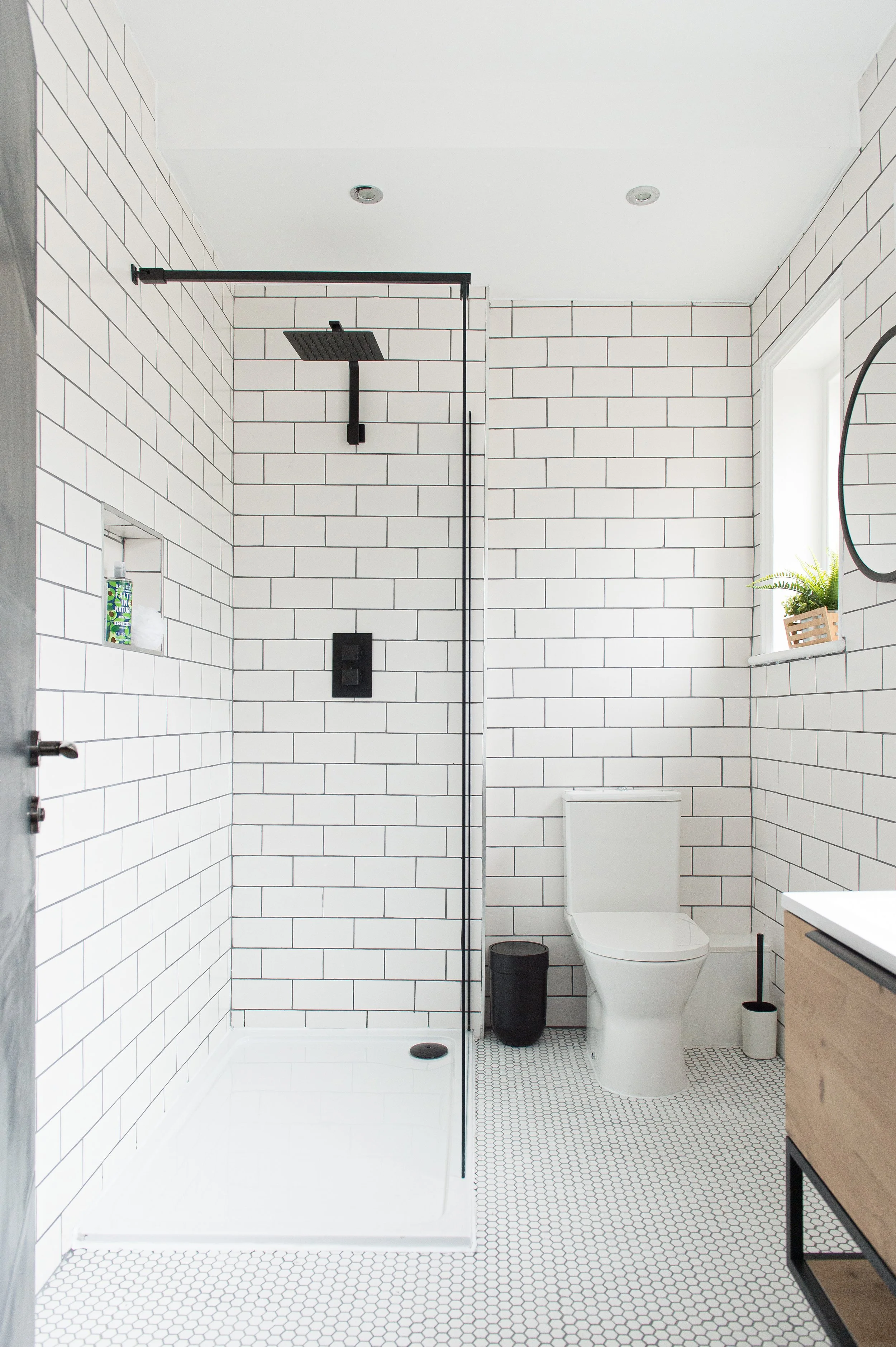 Modern bathroom with white subway tile walls, a walk-in shower with black fixtures, and a white toilet. The floor has small hexagonal tiles, and there is a window with a small plant on the sill.
